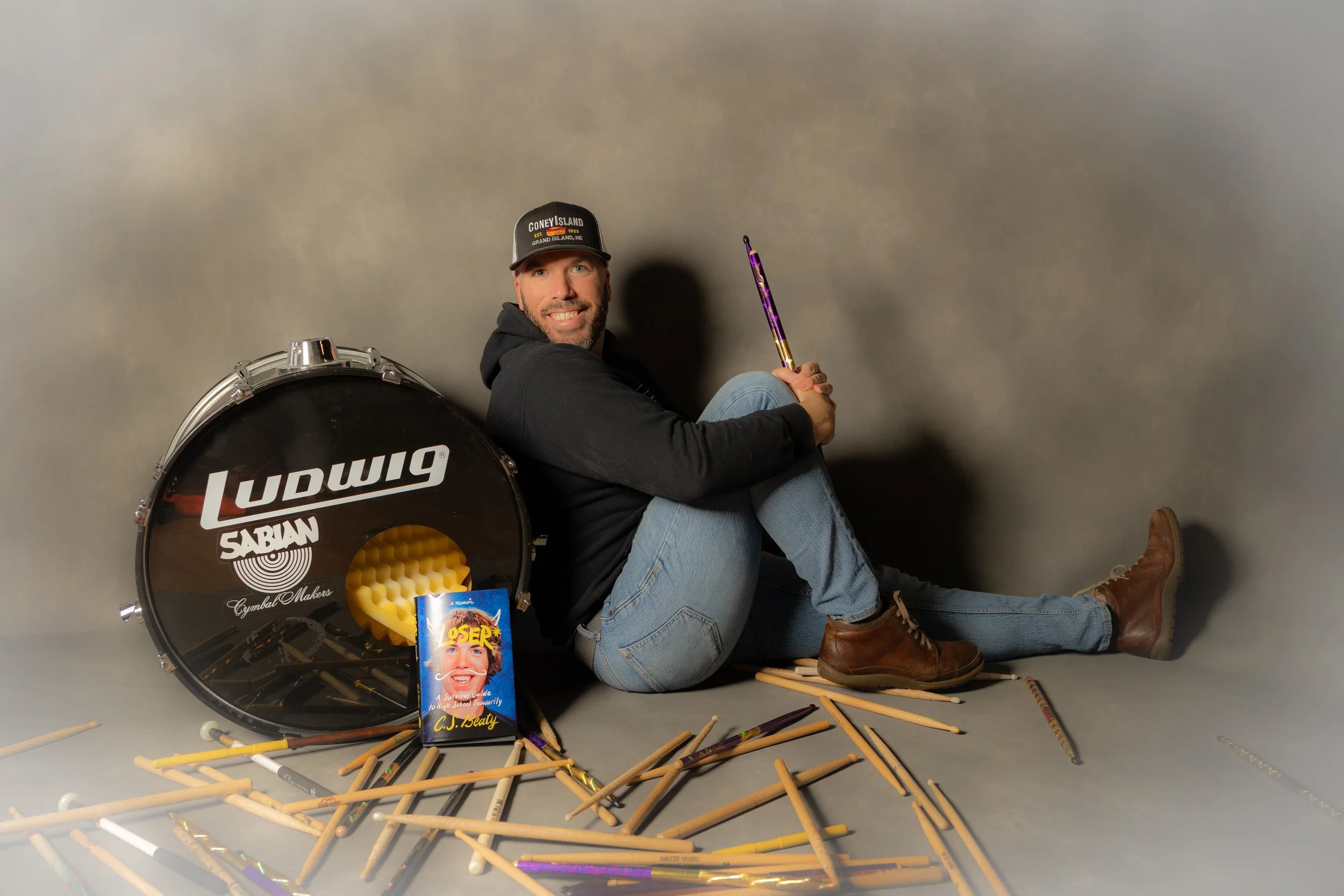 A man sitting on the floor, leaning against a bass drum with a Ludwig logo, surrounded by drumsticks, a book, and percussion equipment, holding a colorful object, wearing a black hoodie, jeans, brown boots, and a cap.