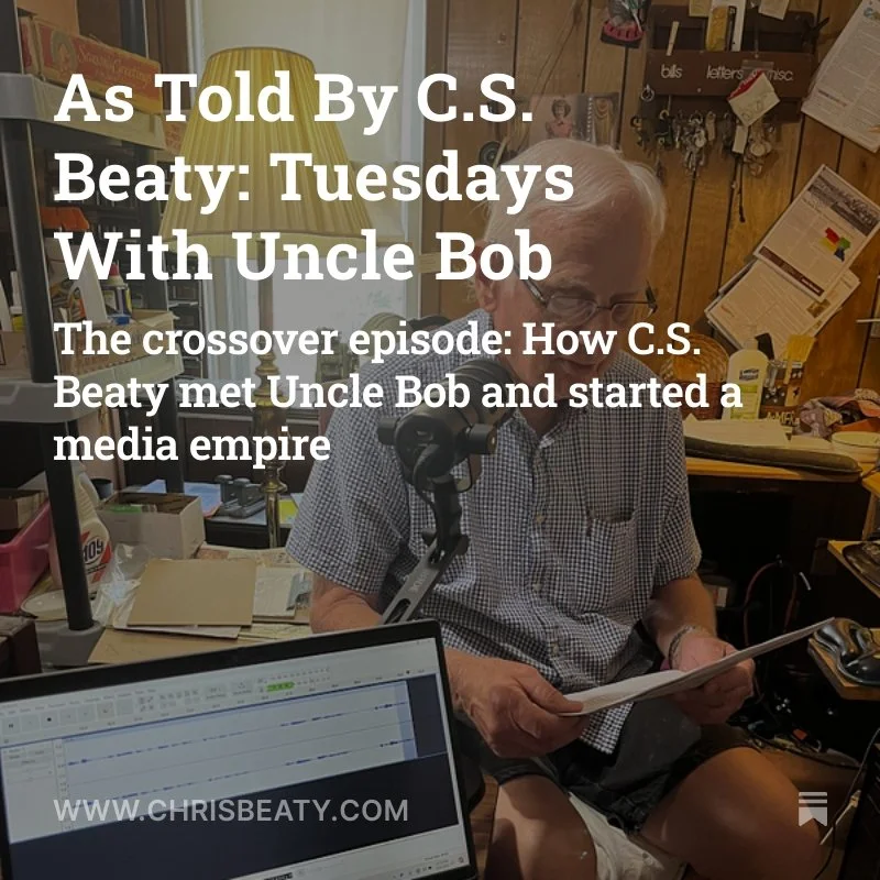 An elderly man with white hair and glasses, wearing a plaid short-sleeve shirt, sitting at a cluttered desk with a microphone, reading a piece of paper, with various papers, books, and a computer screen in the background.