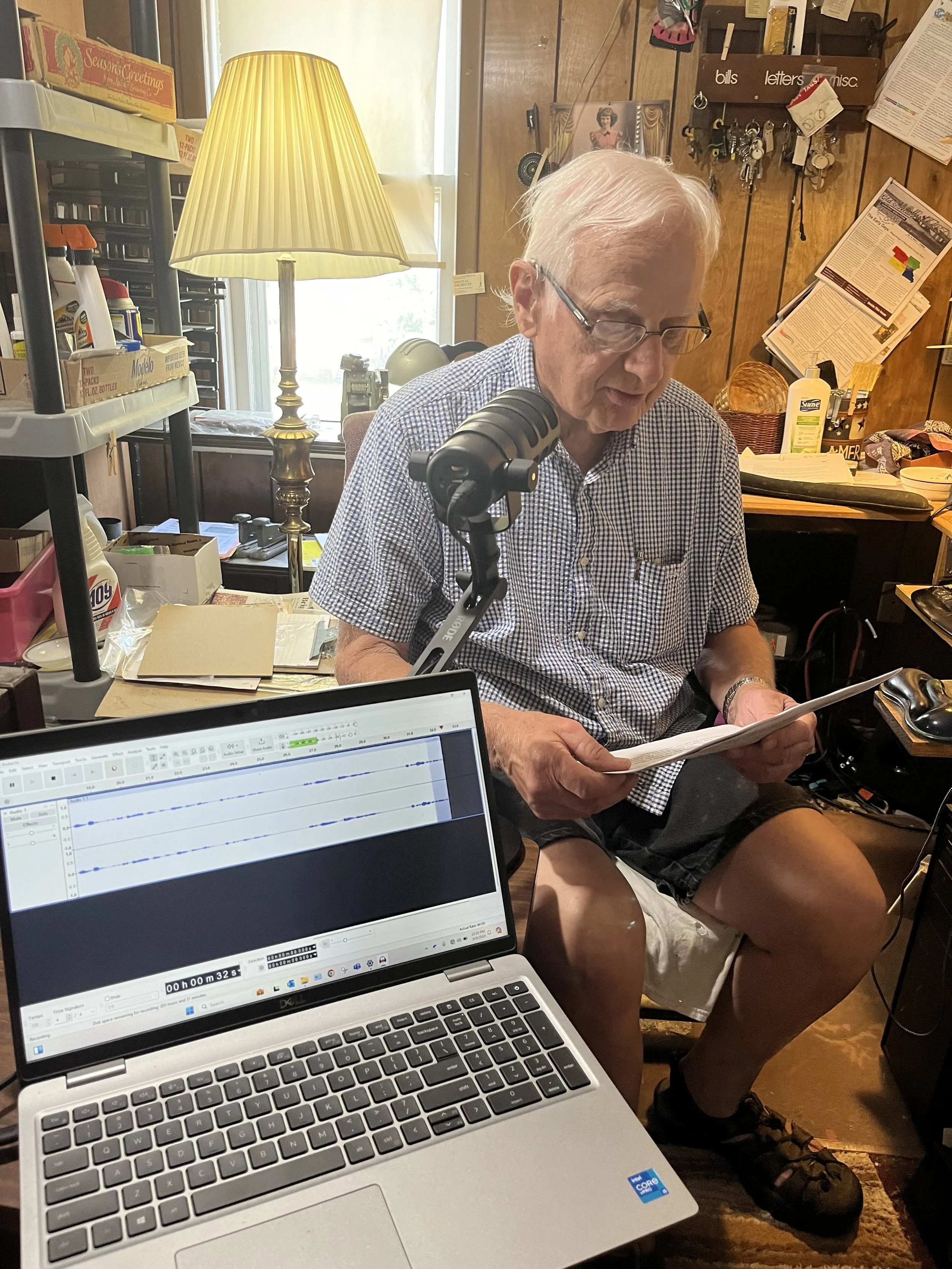 An elderly man seated in a cluttered workspace, reading a paper, with a microphone in front of him, a laptop open with audio editing software, a standing lamp, and various office supplies and posters on the wooden paneled wall behind him.