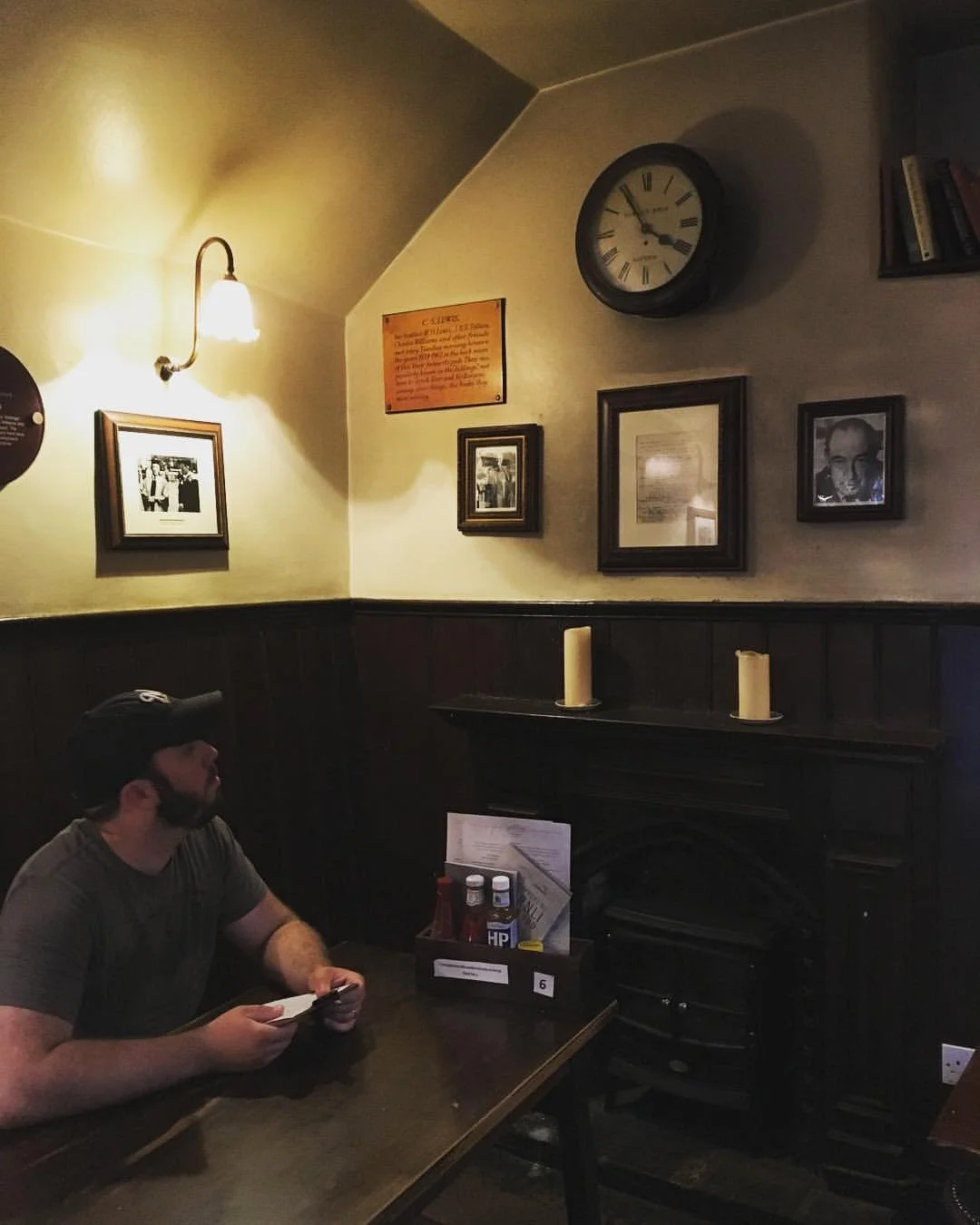 A man sitting at a table in a dimly lit restaurant or pub, holding a phone in one hand. The wall behind him has framed photos, a clock, and candles. There is a wall-mounted lamp on the wall.