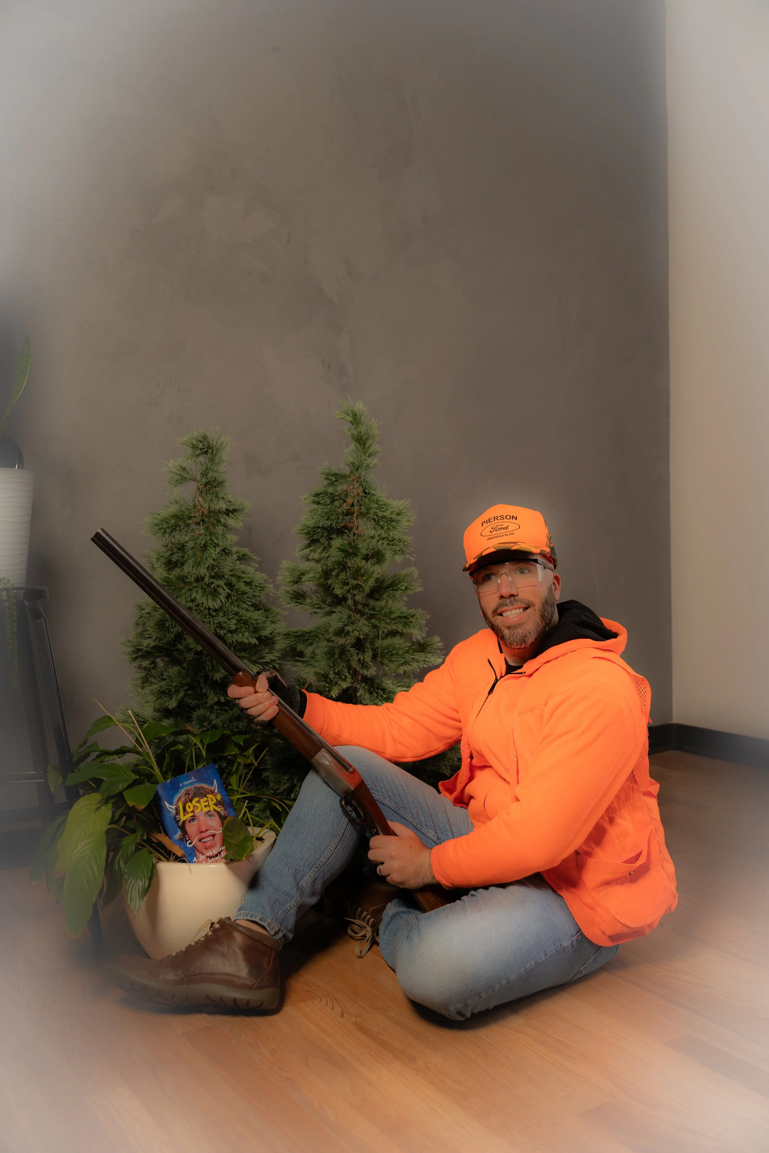 A man in an orange jacket and cap sitting on the floor with a rifle, smiling at the camera, next to two small Christmas trees and a potted plant with a humorous magazine titled 'Loser' behind it.