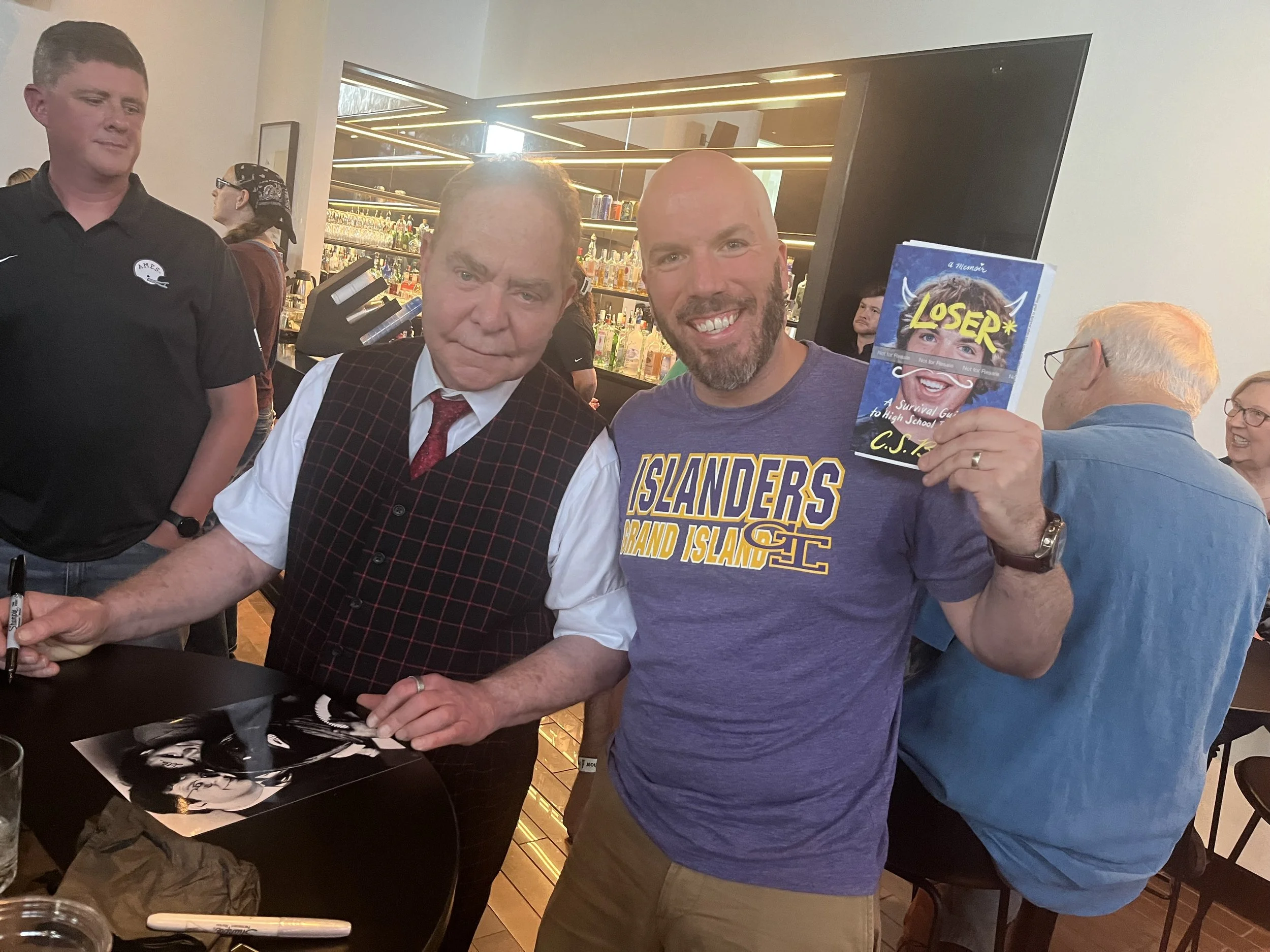 A man holding a signed photograph and a book titled "LOSER" at a book signing event, standing next to a man in a vest signing the photograph, with other people in the background in a bar or restaurant setting.