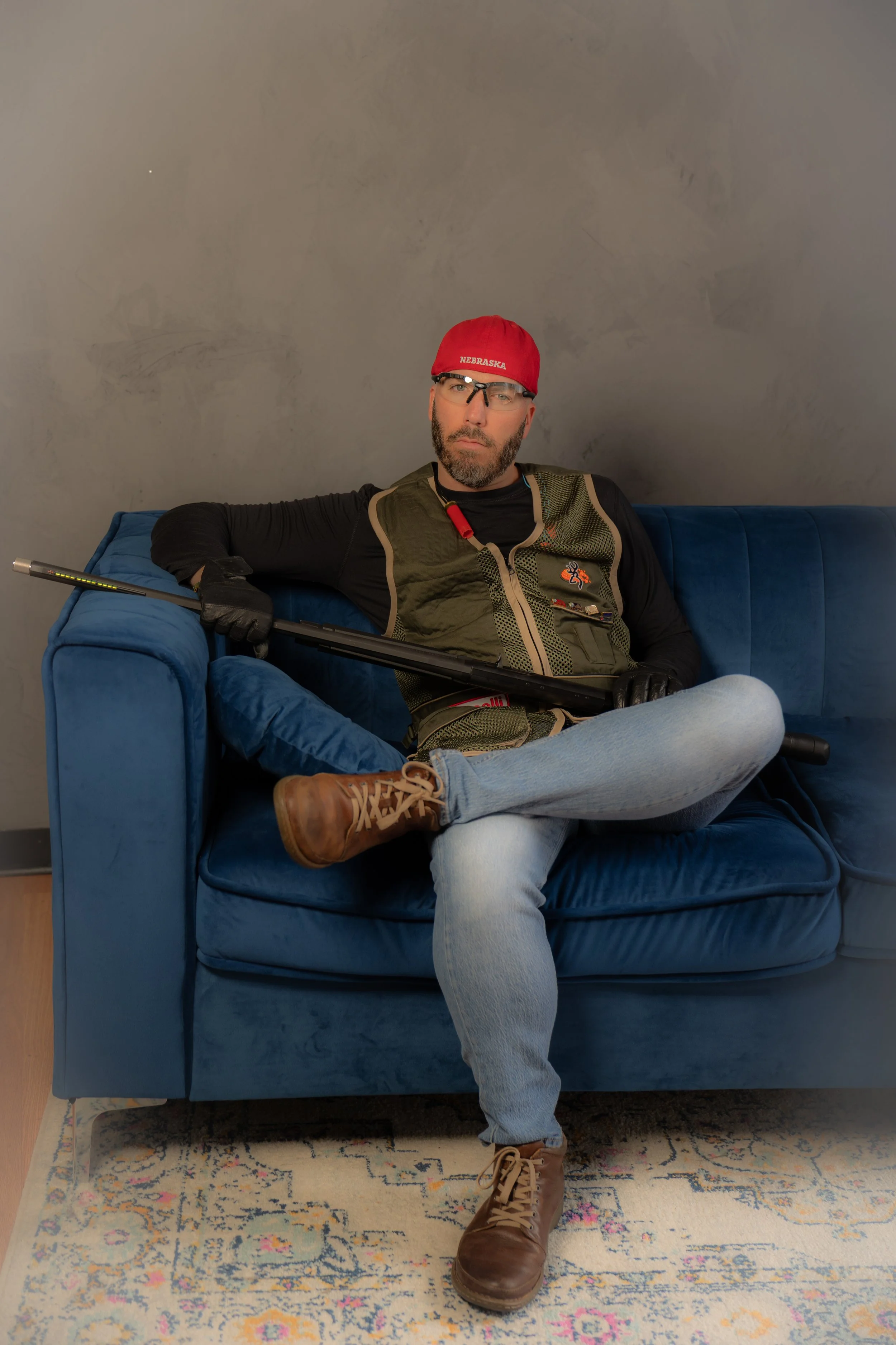 A man with a beard and glasses sitting on a blue sofa, wearing a red cap, black shirt, vest, jeans, and brown boots, holding a golf club.