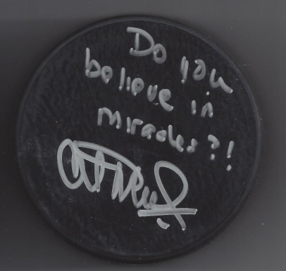 A circular black sign with white handwritten text that reads, 'Do you do love in miracles?!' and a signature at the bottom.