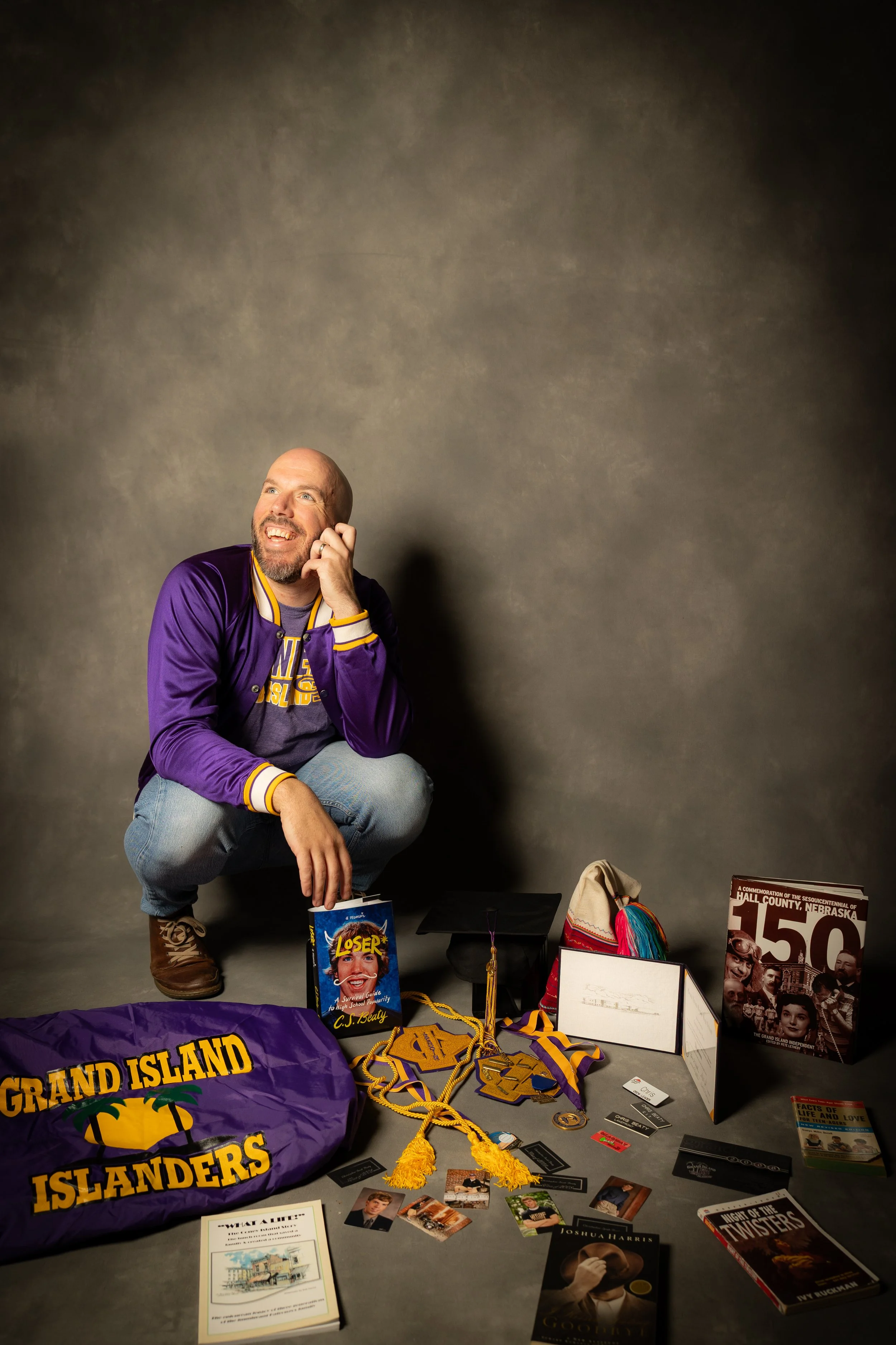 A man crouching down, smiling, wearing a purple and yellow Grand Island Islanders jacket, surrounded by various books, medals, photographs, a graduation cap, and memorabilia related to Grand Island, Nebraska.