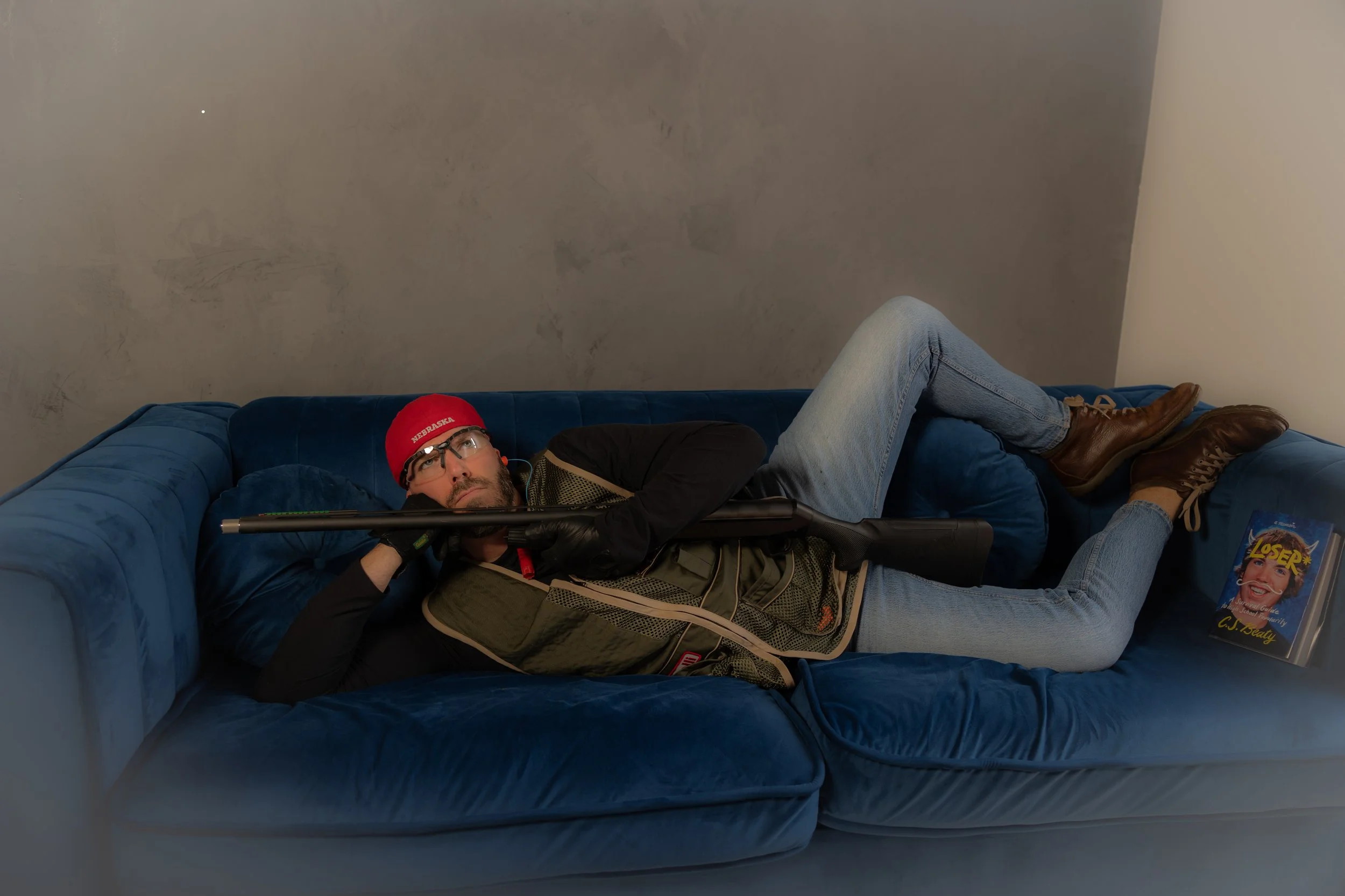 Man lying on a blue couch, wearing glasses, a red beanie, and holding a rifle, with a magazine titled 'Loser' on the floor beside him.