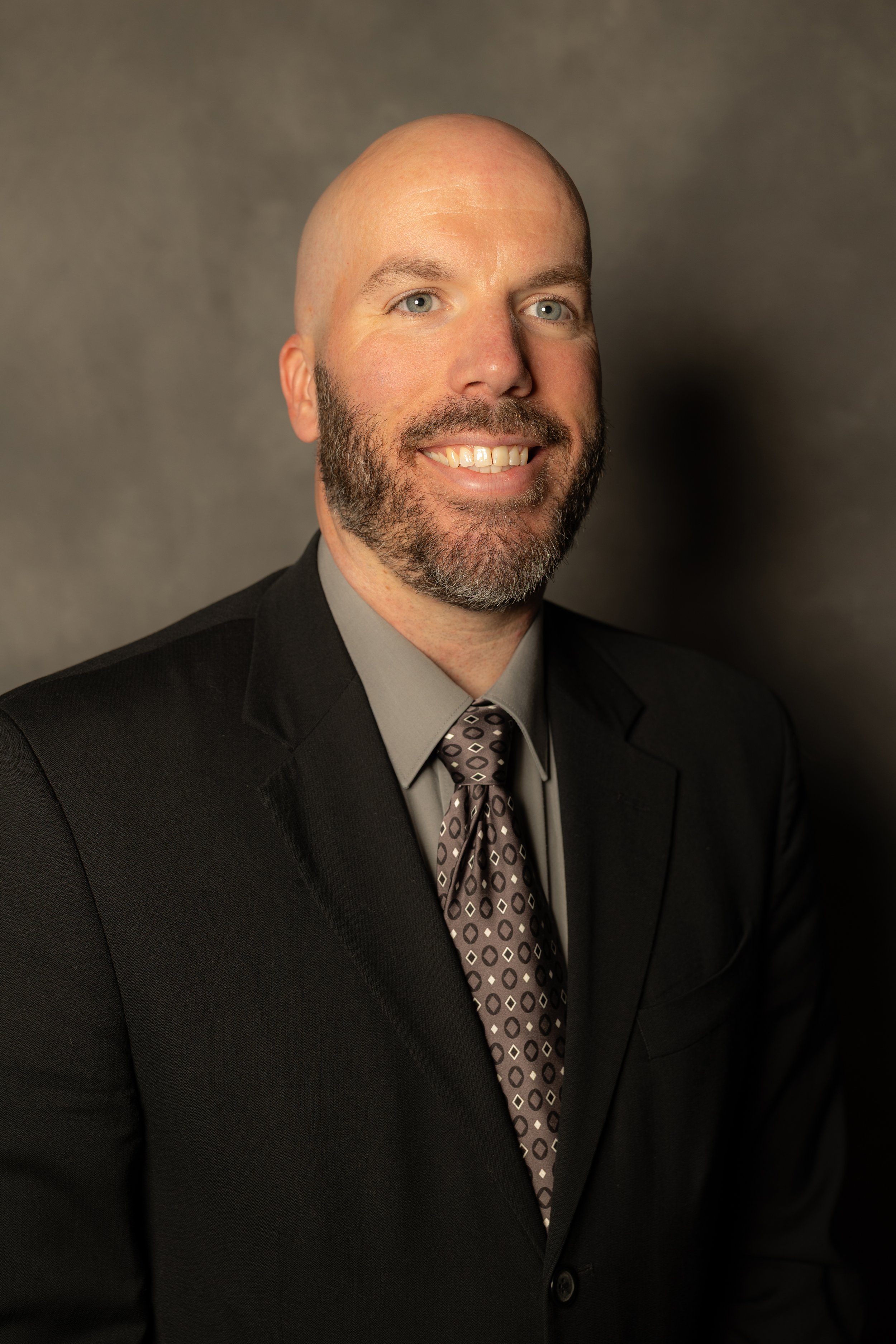 A professional man with a beard and bald head, wearing a black suit, gray shirt, and patterned tie, smiling in front of a dark gray background.