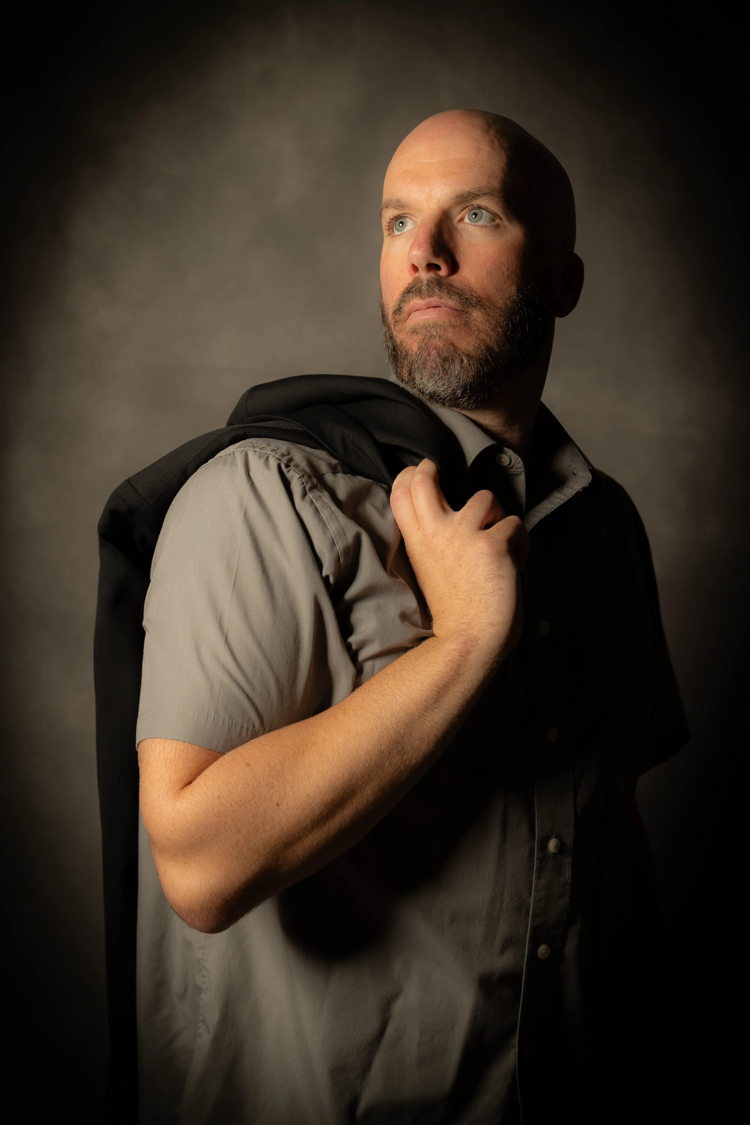 A man with a bald head and a beard holds a black jacket over his shoulder, looking upward against a dark background.