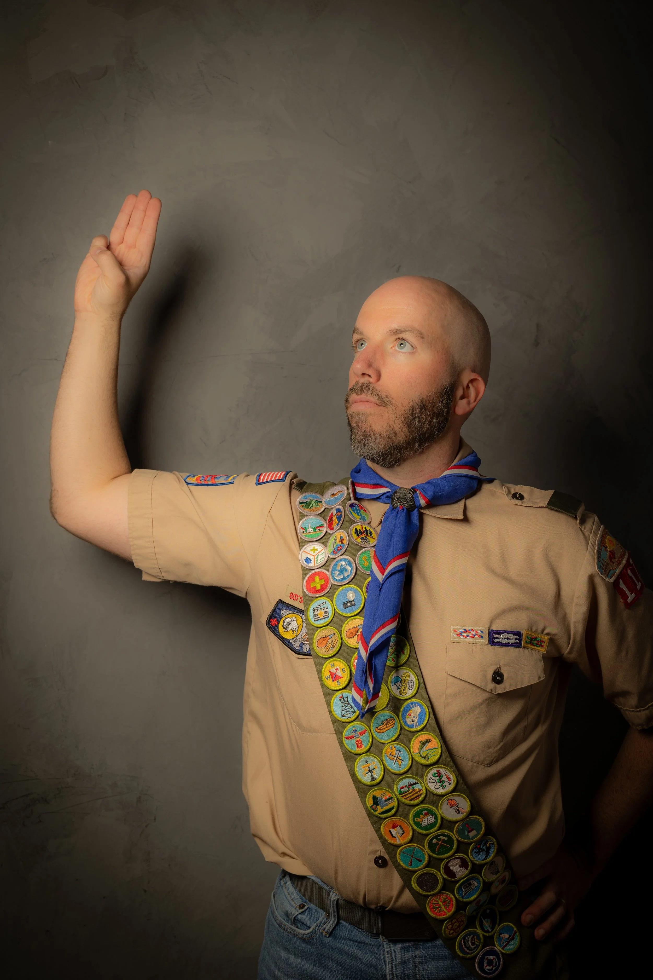 A man in a Boy Scout uniform with numerous badges and a neckerchief, raising his right hand in a scout salute against a dark background.