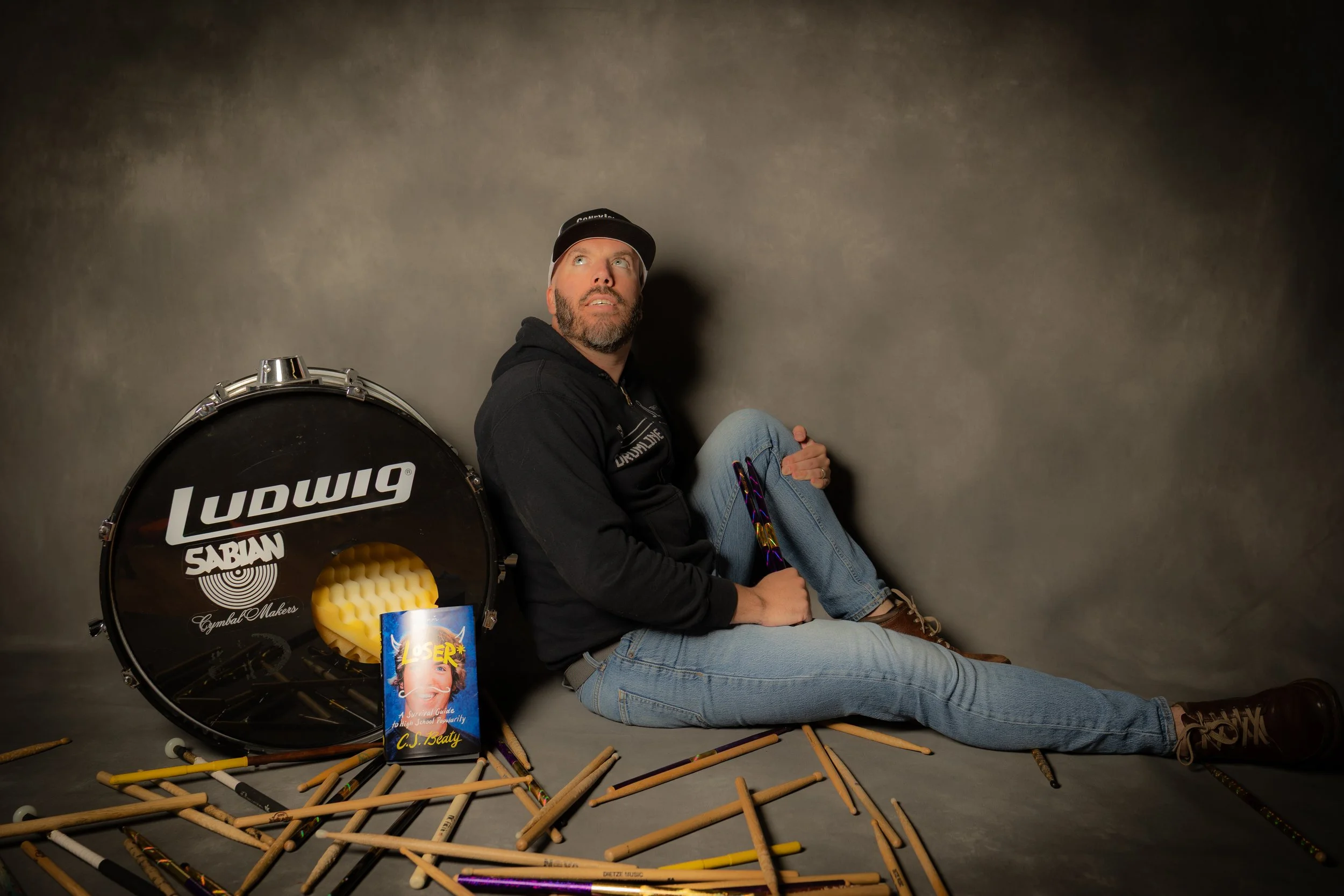 A man with a beard and gray hair, wearing a black hoodie, jeans, and brown boots, sitting on the floor against a gray backdrop. He is surrounded by drumsticks, a Ludwig Snare drum, a book, and some colorful objects, looking up and slightly to the sid
