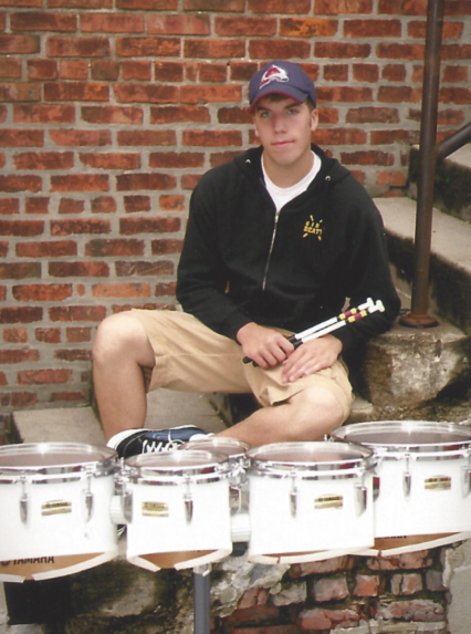 Young man sitting outdoors in front of a brick wall, holding drumsticks, with a set of drums in front of him.