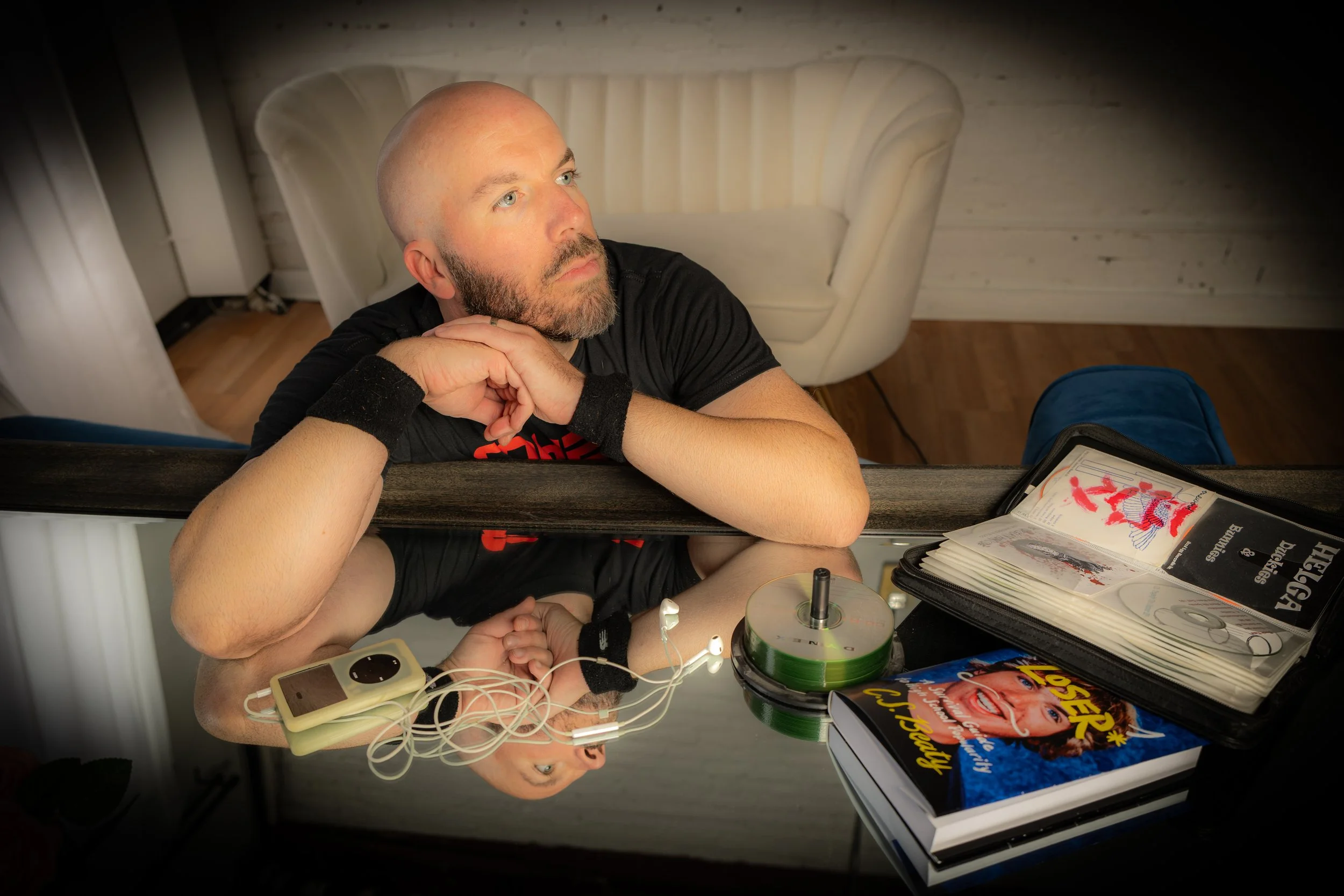 A bald man with a beard is resting his chin on his hand, looking up thoughtfully. He is sitting at a table with a mirror, on which his reflection and various books and a CD are visible. The room has a white couch and wooden floor in the background.