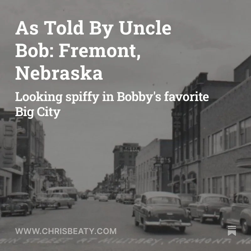 Black and white photo of a downtown street scene with vintage cars parked along the sidewalk and buildings on either side.