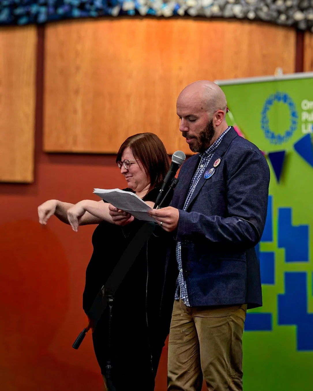 A man and woman on stage at a public event. The man is speaking into a microphone and holding papers, while the woman is signing or gesturing with her hands. A green and blue banner is in the background.