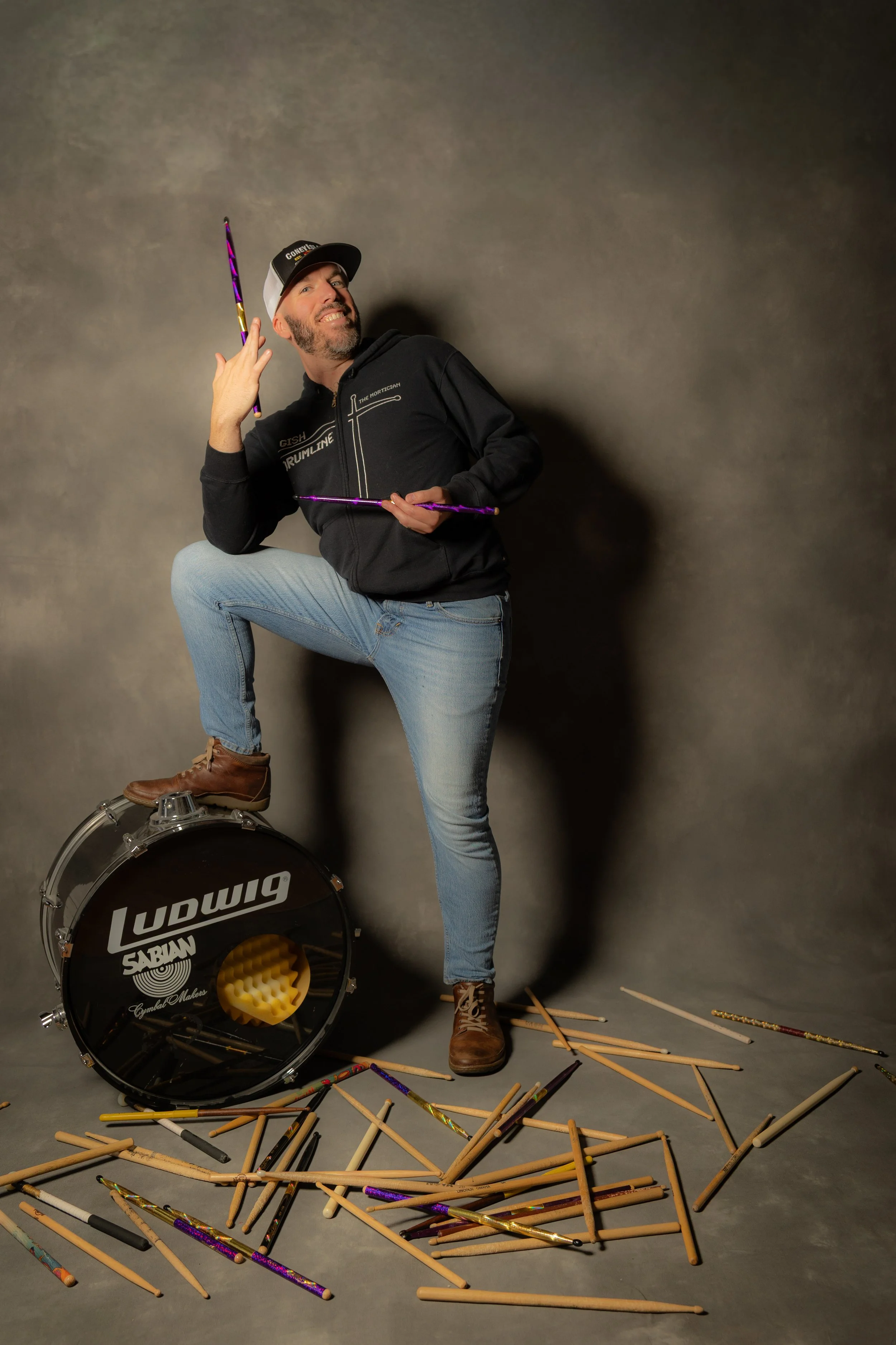 A man with a beard and mustache is posing dramatically, holding drumsticks, with his right foot on a Ludwig bass drum, while surrounded by scattered drumsticks, on a gray studio background.