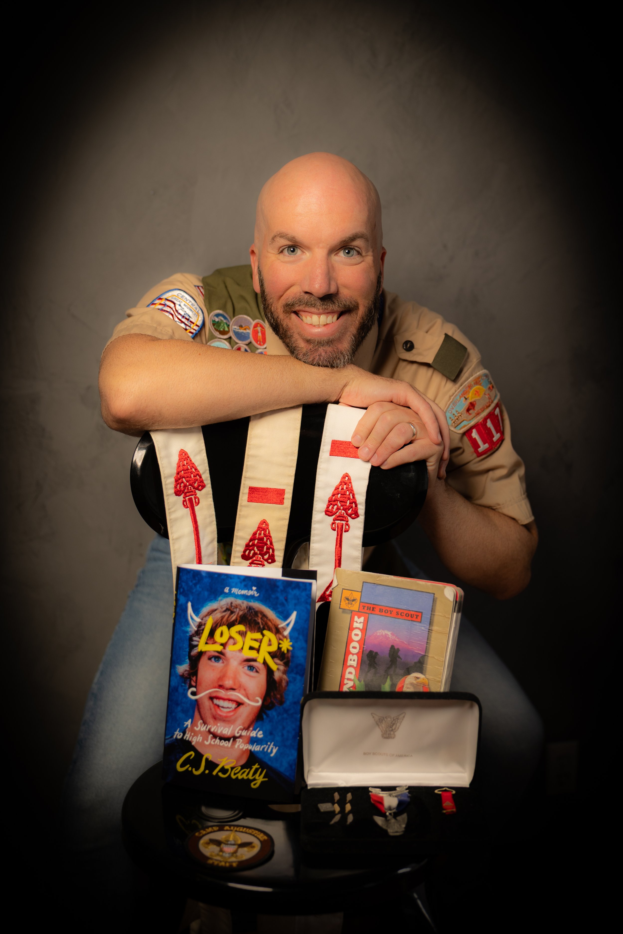 A smiling man in a Boy Scouts uniform leaning on a chair with books, badges, and medals on display.