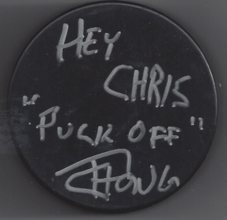 A round black hockey puck with handwritten silver text saying, "Hey Chris, puck off!" signed by an illegible signature.