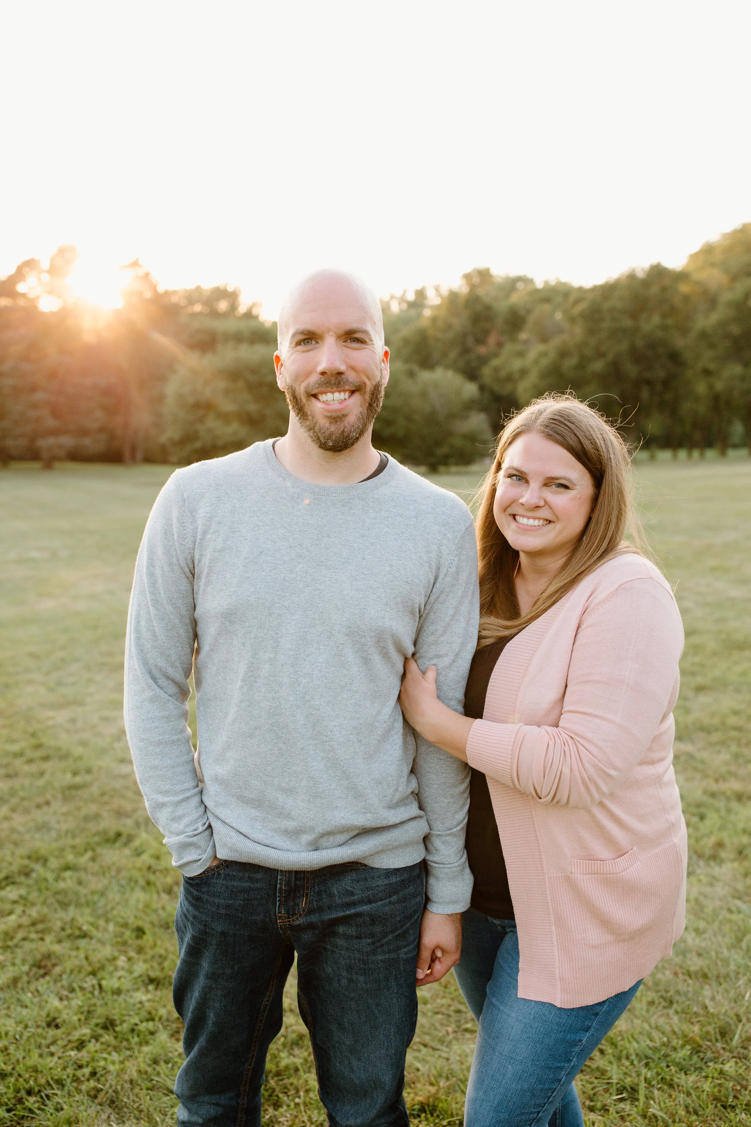 A smiling couple standing on a grassy field during sunset, with trees in the background.