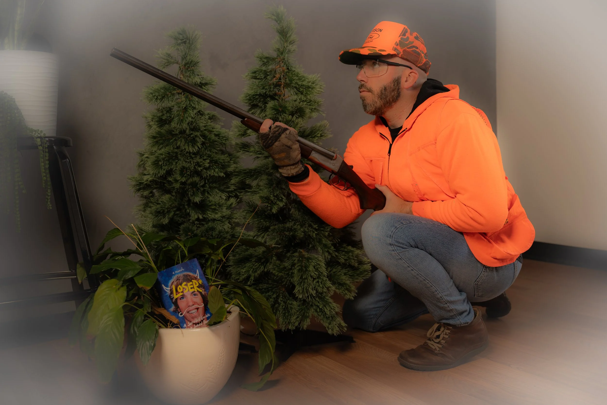 A man dressed in an orange jacket, glasses, and a cap is kneeling down, aiming a shotgun at a small artificial Christmas tree. There is a potted houseplant with a parody magazine cover titled 'LOSER' in the background.