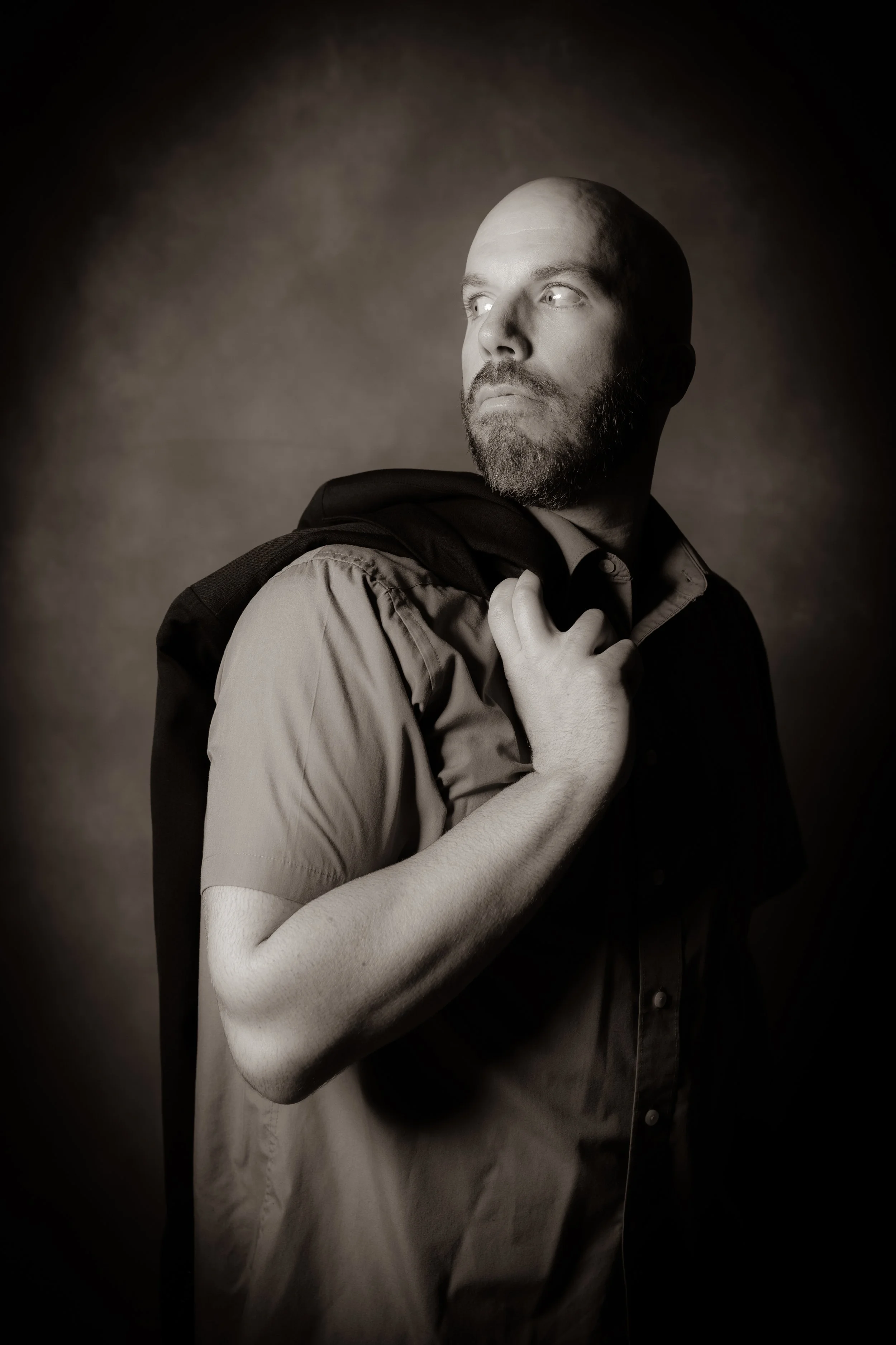 Black and white portrait of a man with a beard, wearing a short-sleeved shirt, holding a jacket over his shoulder, looking to the side.
