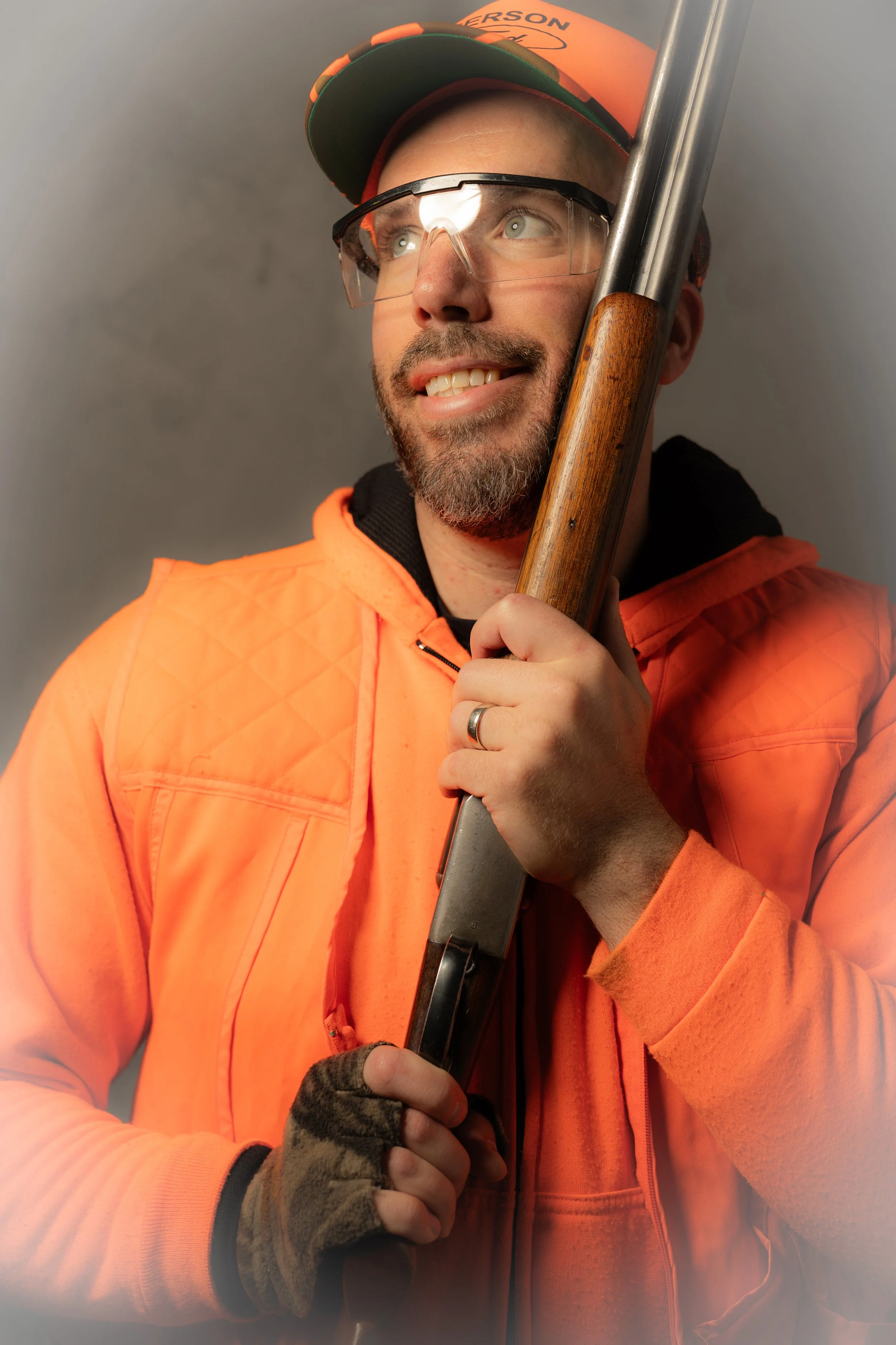 A man in safety glasses, an orange cap, and an orange jacket holding a shotgun, with a smile and looking upward.