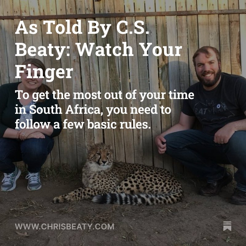 Two people sitting beside a leopard cub in front of a wooden fence. The person on the left is wearing glasses, a black top, and jeans with sneakers, while the person on the right has a beard, is smiling, and is wearing a dark T-shirt and pants. The text overlay reads: "As Told By C.S. Beaty: Watch Your Finger. To get the most out of your time in South Africa, you need to follow a few basic rules." The website www.chrisbeaty.com is at the bottom of the image.