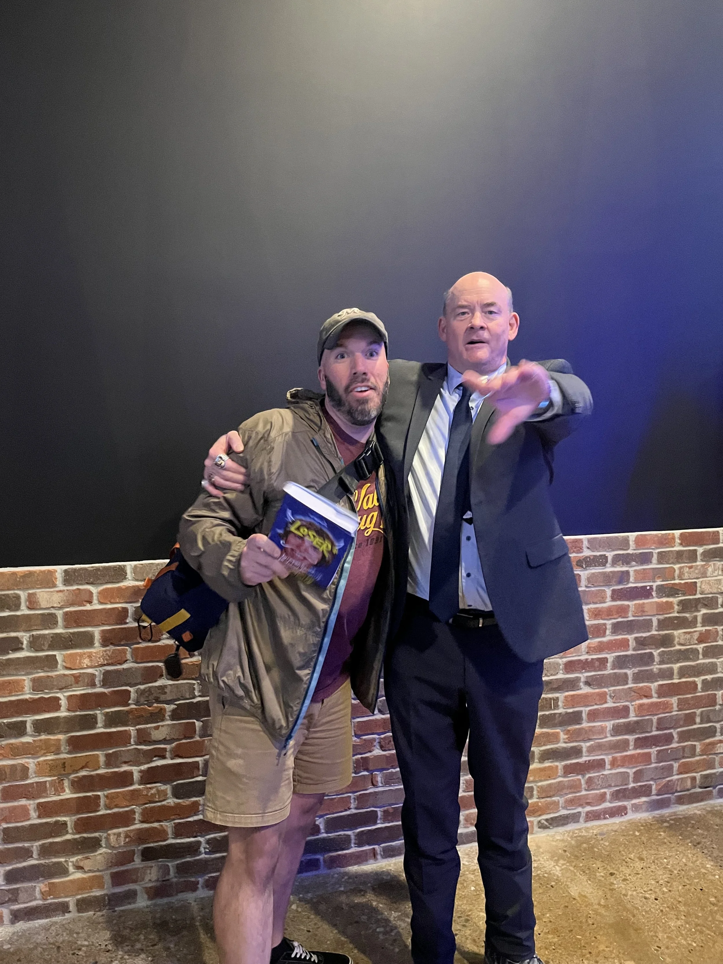 Two men posing together, one holding a snack bag and backpack, the other gesturing with his hand, standing against a brick wall.