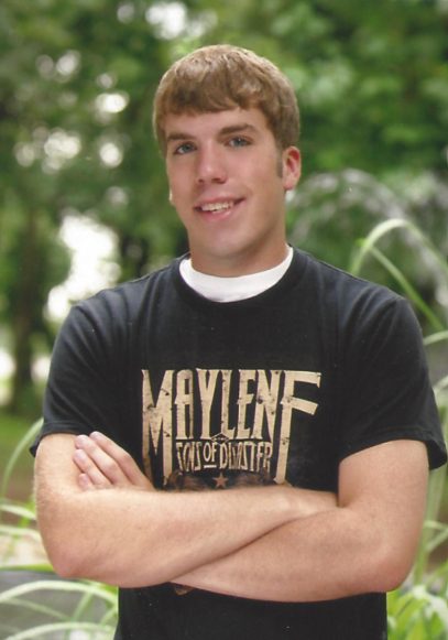 Young man with short brown hair wearing a black T-shirt with a Maylene band logo, standing outdoors with greenery in the background.
