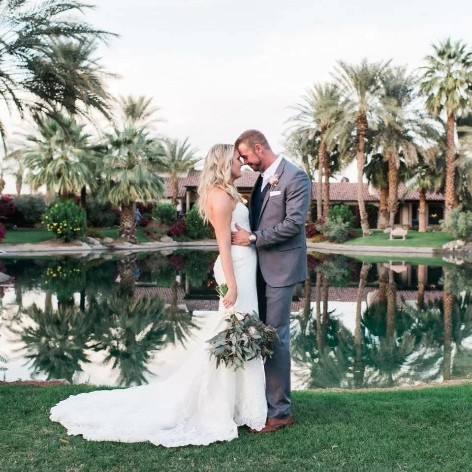 A newlywed couple in wedding attire standing close to each other by a pond with reflections, surrounded by palm trees and lush greenery.