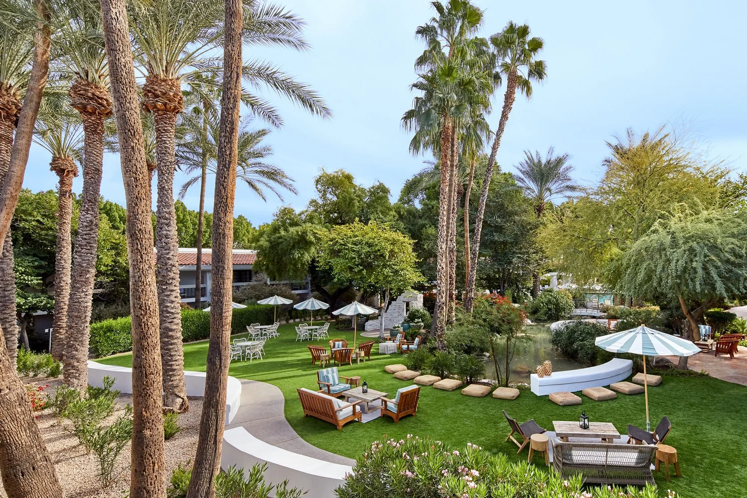 An outdoor garden area with lush green grass, large palm trees, various seating options like wooden benches, cushioned chairs, and tables with umbrellas, surrounded by trees and shrubs, under a clear blue sky.