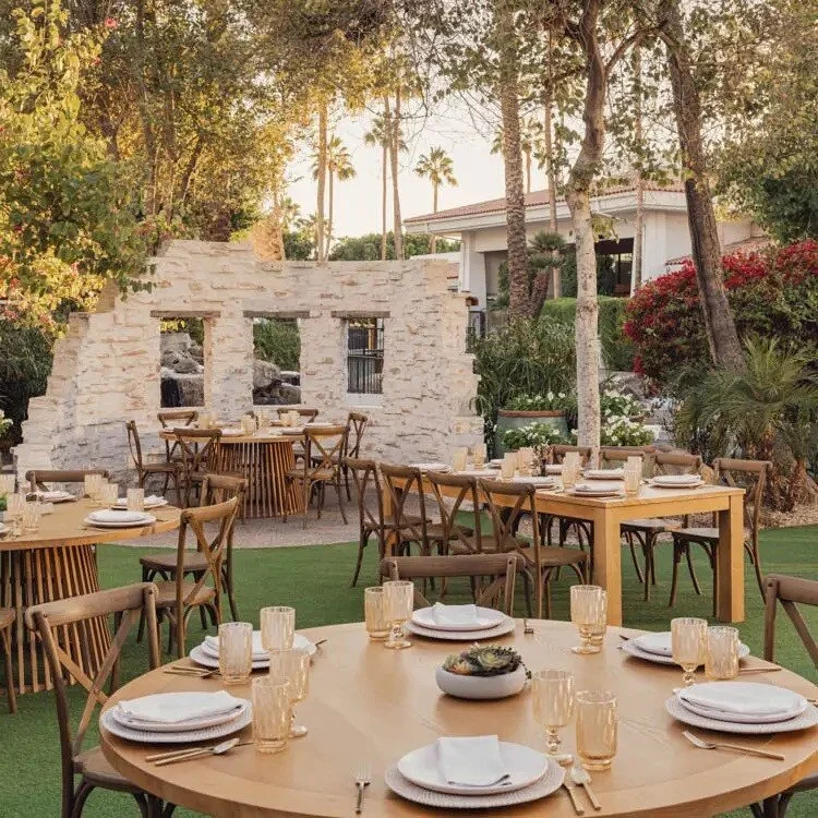 Outdoor dining setup with round and rectangular wooden tables, chairs, white plates, napkins, and beige glasses, surrounded by trees and a stone wall in the background.