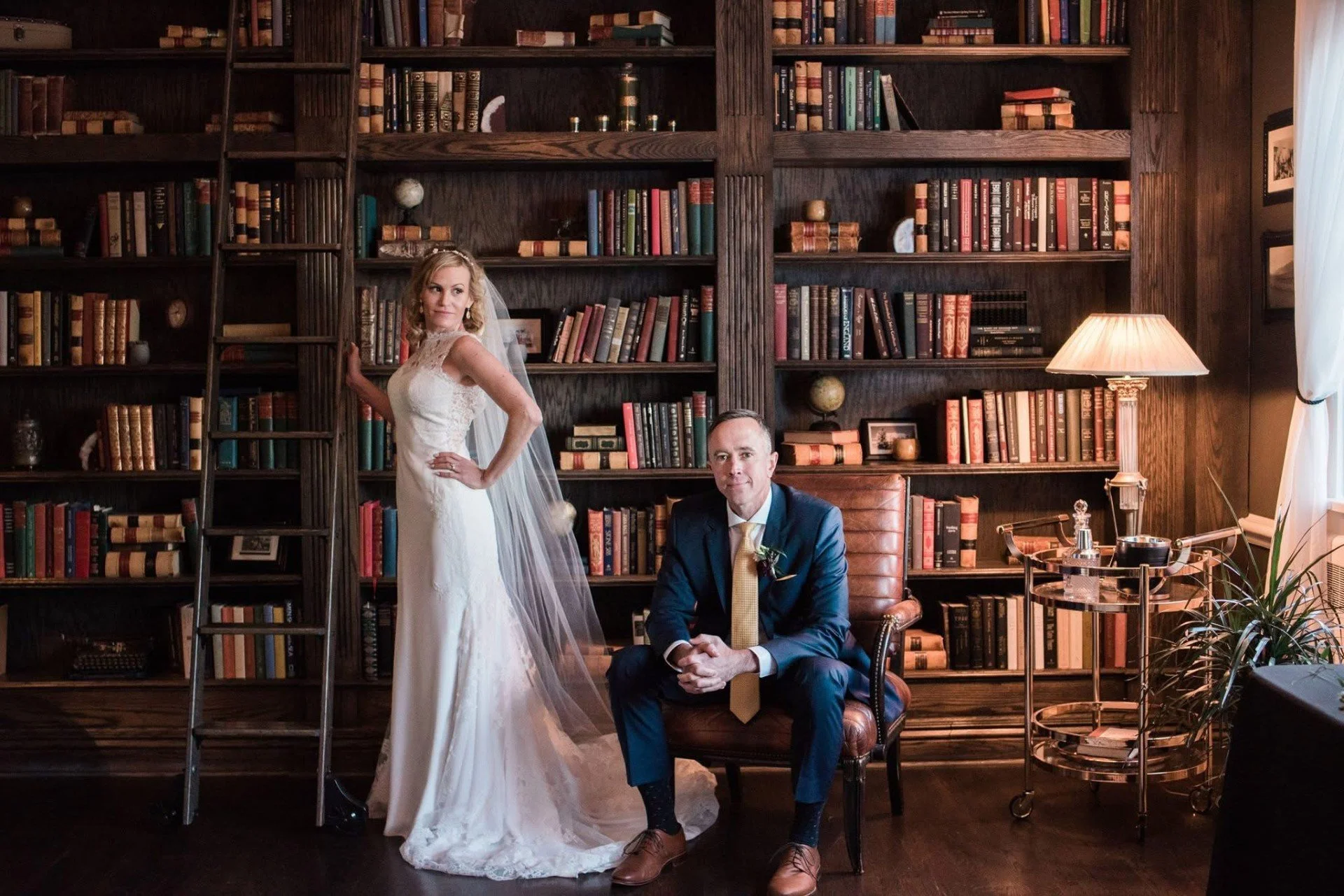 A bride in a white wedding dress standing next to a groom in a suit, seated, in front of a large dark wooden bookshelf filled with books, in a cozy, well-lit room.