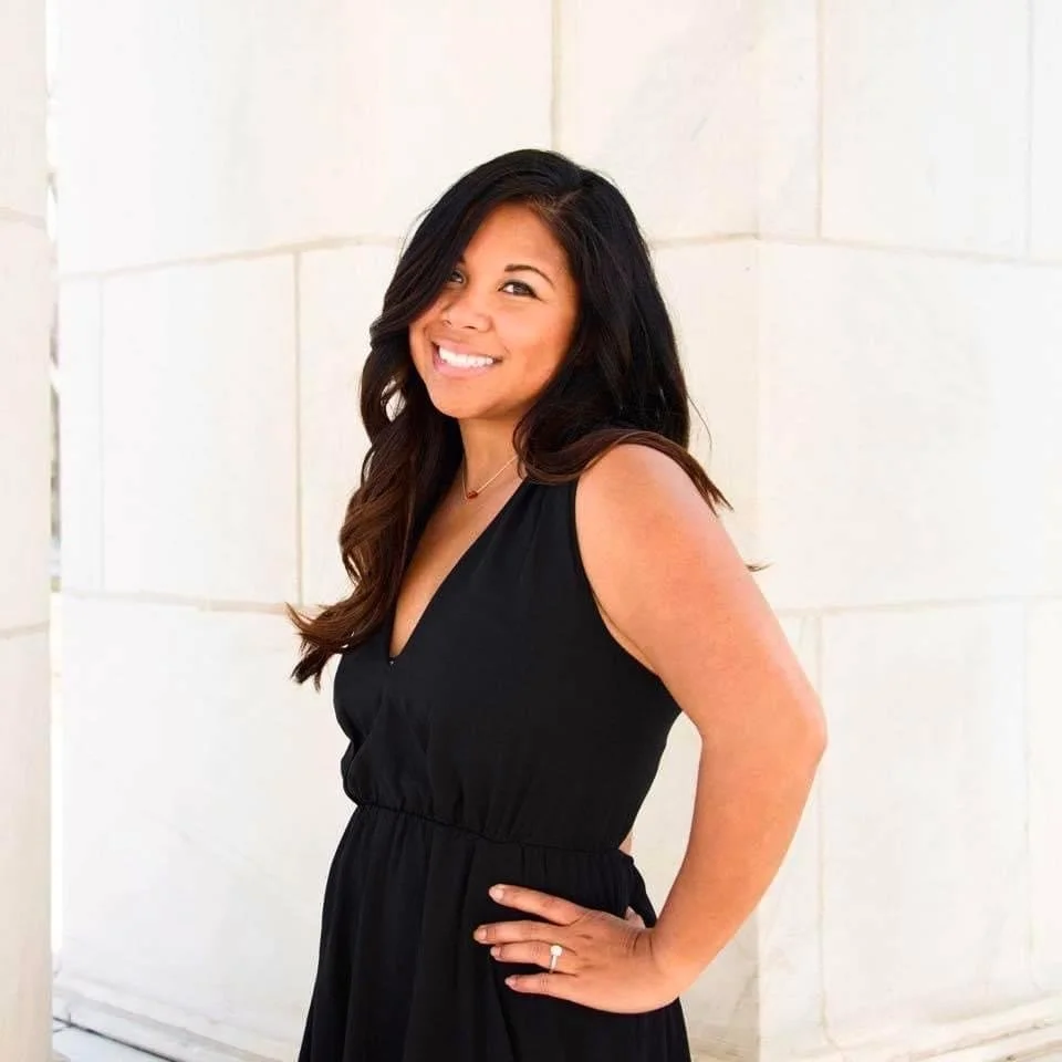 A woman with long dark hair, smiling, wearing a sleeveless black dress, standing against a light-colored stone wall.