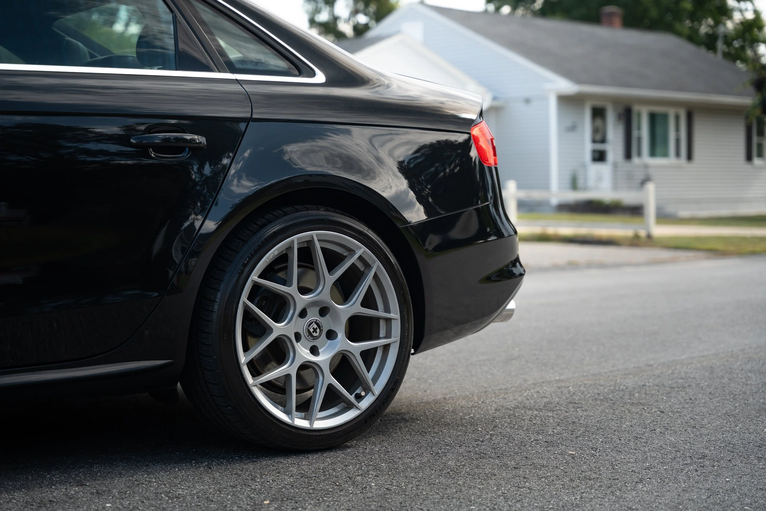 Close-up of the rear side of a black luxury sedan car parked on a residential street, showing the rear wheel and part of the trunk, with a house in the background.