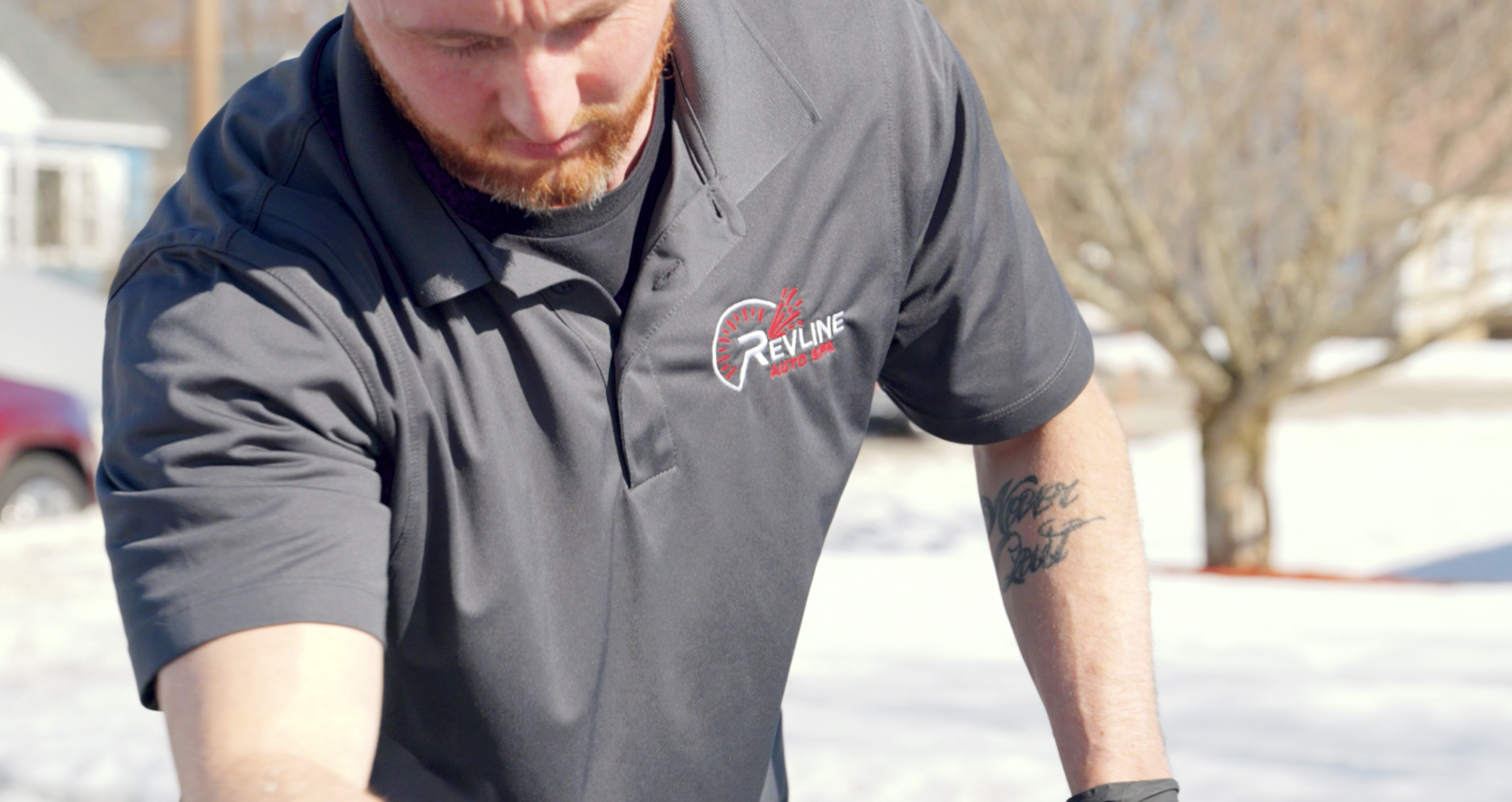Man wearing a grey polo shirt with a logo that says "Revline Auto Spa" on the chest, outdoors on a snowy day, with a tree and cars in the background.