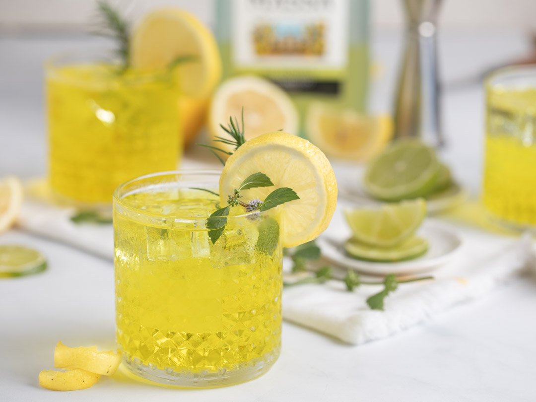 A glass of lemonade with lemon slices and mint leaves, with additional lemons, limes, and bottles of lemonade in the background.