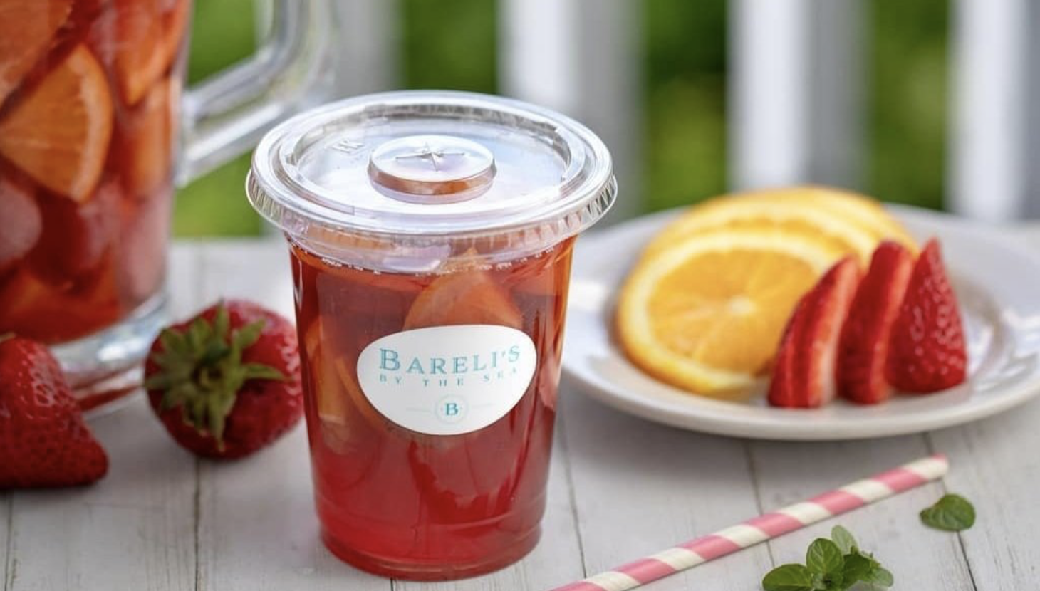 Clear plastic cup of iced tea with orange slices, with strawberries and sliced oranges and strawberries on a plate in the background.