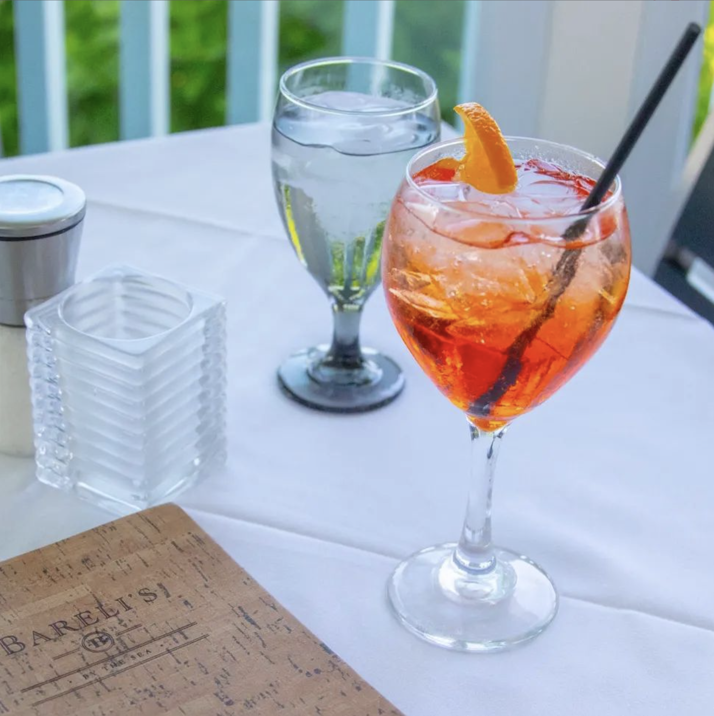A table with a glass of water, a list, a stack of napkins or coasters, and a large glass of Aperol spritz with an orange slice and a straw, set outdoors with green foliage in the background.