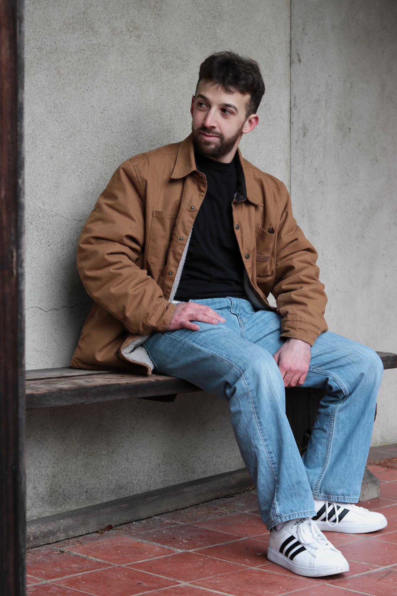 A young man sitting on a wooden bench against a concrete wall, wearing a brown jacket, black shirt, light blue jeans, and white sneakers with black stripes.