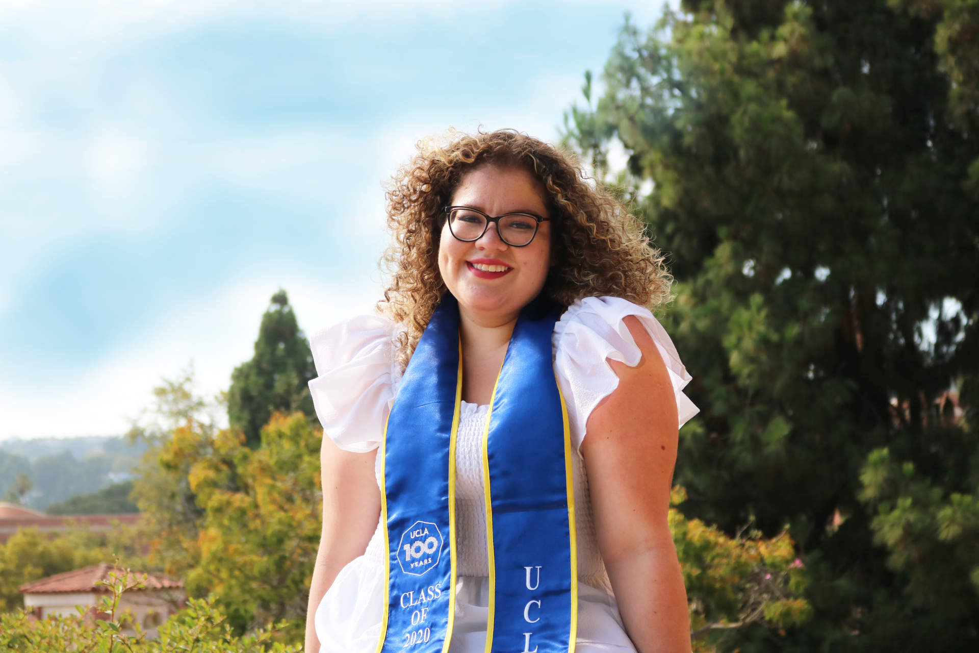A woman with curly hair and glasses, wearing a white dress and a blue class of 2020 sash, stands outdoors with trees and a clear sky in the background, smiling at the camera.