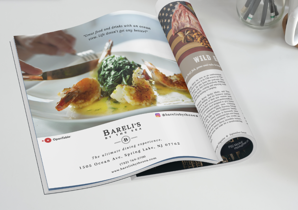 Open magazine with a photo of a seafood dish on the page, featuring shrimp, greens, and sauce, over a white table. Part of another page is curled up, showing text and images. The restaurant name is 'Bareli's by the Sea'.