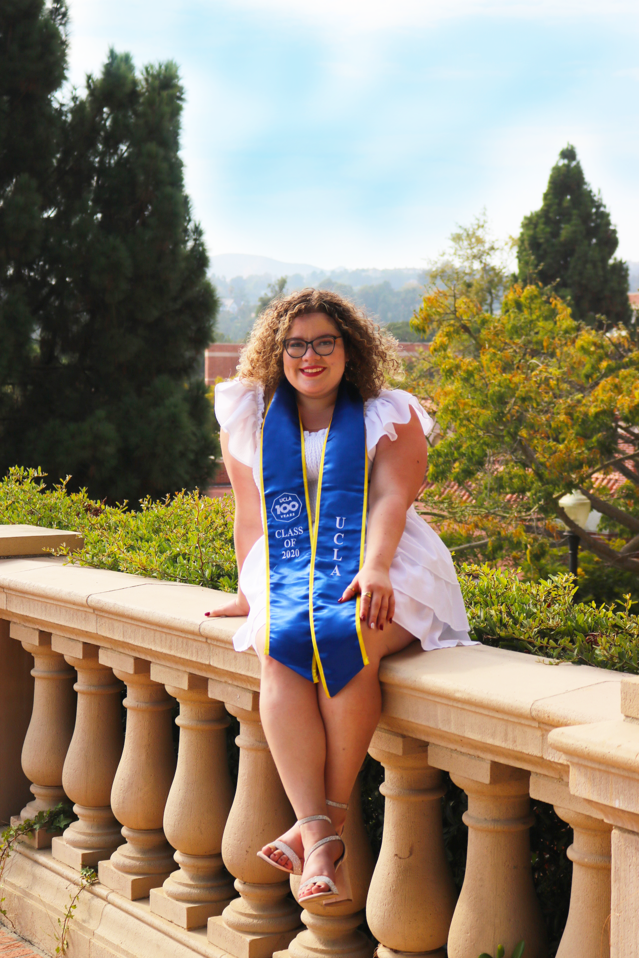 Young woman with curly hair wearing glasses, a white dress, and a blue graduation stole with yellow trim, sitting on a stone railing outdoors. She has a big smile, dark lipstick, and is wearing silver sandals. The background features trees and a clea