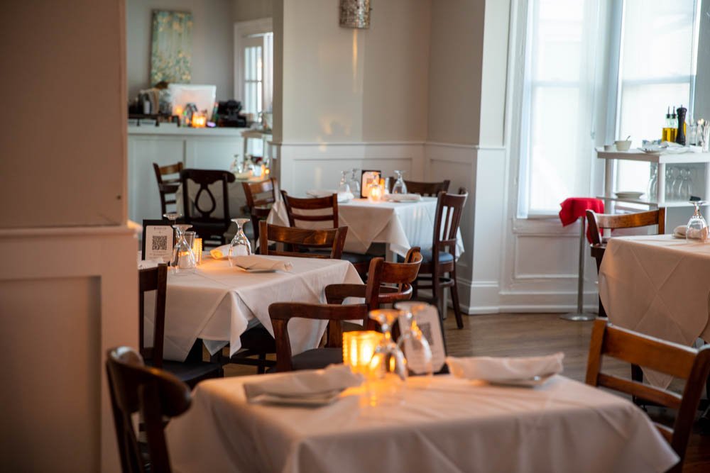 Empty dining room with tables set for service, white tablecloths, wine glasses, and candles, in a softly lit restaurant with windows and natural light.