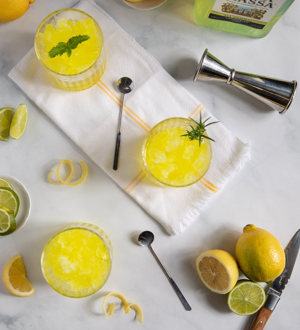 Two lemon cocktails garnished with herbs, surrounded by lemons, lime wedges, lemon peels, a zester, and a muddler, on a white surface.
