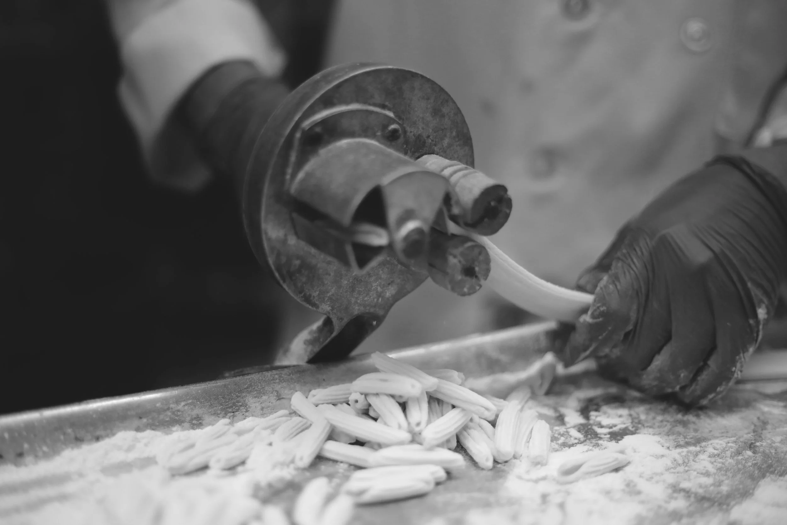A person wearing black gloves uses a pasta machine to roll out pasta dough into noodles.