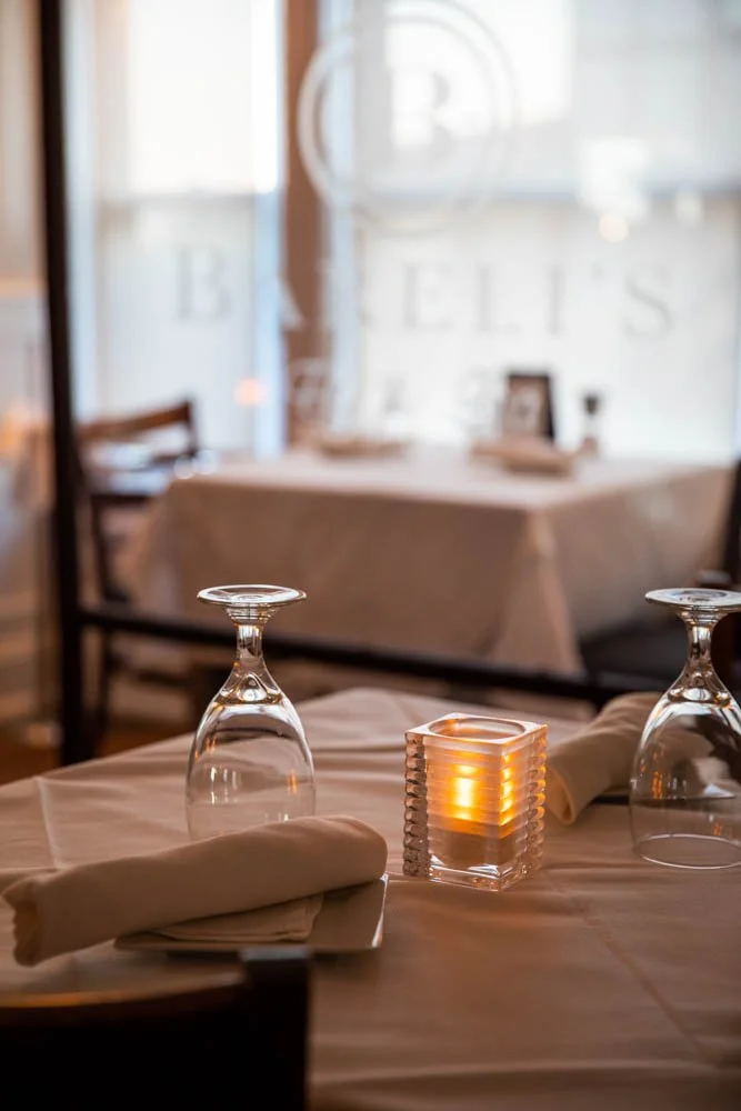 A dining table set with upside-down wine glasses, a folded napkin, and a lit candle in a glass holder in a restaurant with soft lighting and a blurred background featuring a wall with large letters spelling 'BELLS'.