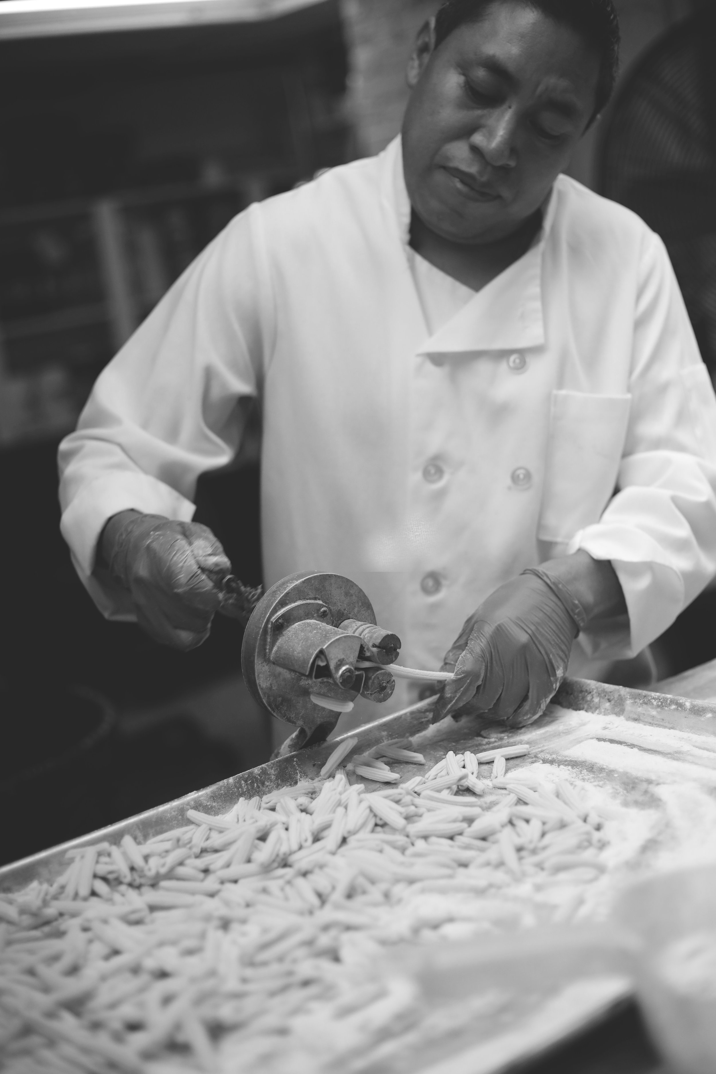 A chef in a white coat and gloves uses a pasta cutter to make pasta on a floured surface.