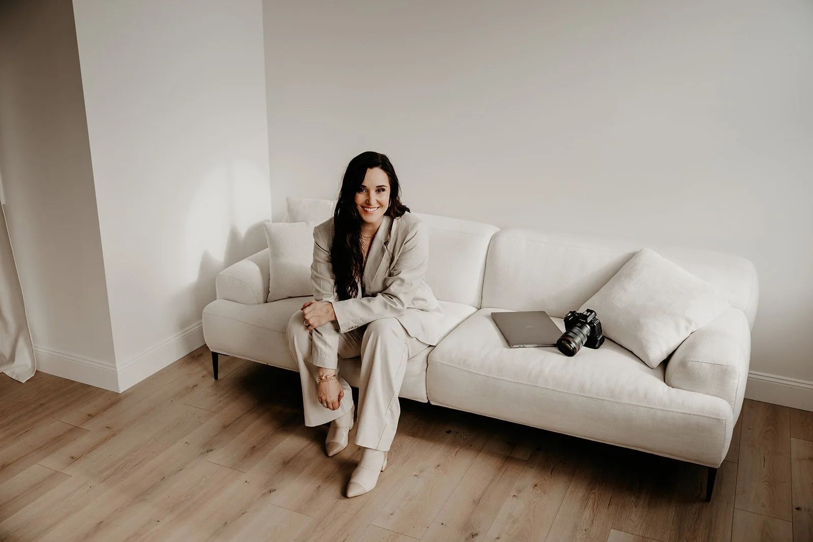 A woman sitting on a white couch in a minimalistic room with wooden floor, smiling at the camera. A camera, a laptop, and a pillow are on the couch beside her.