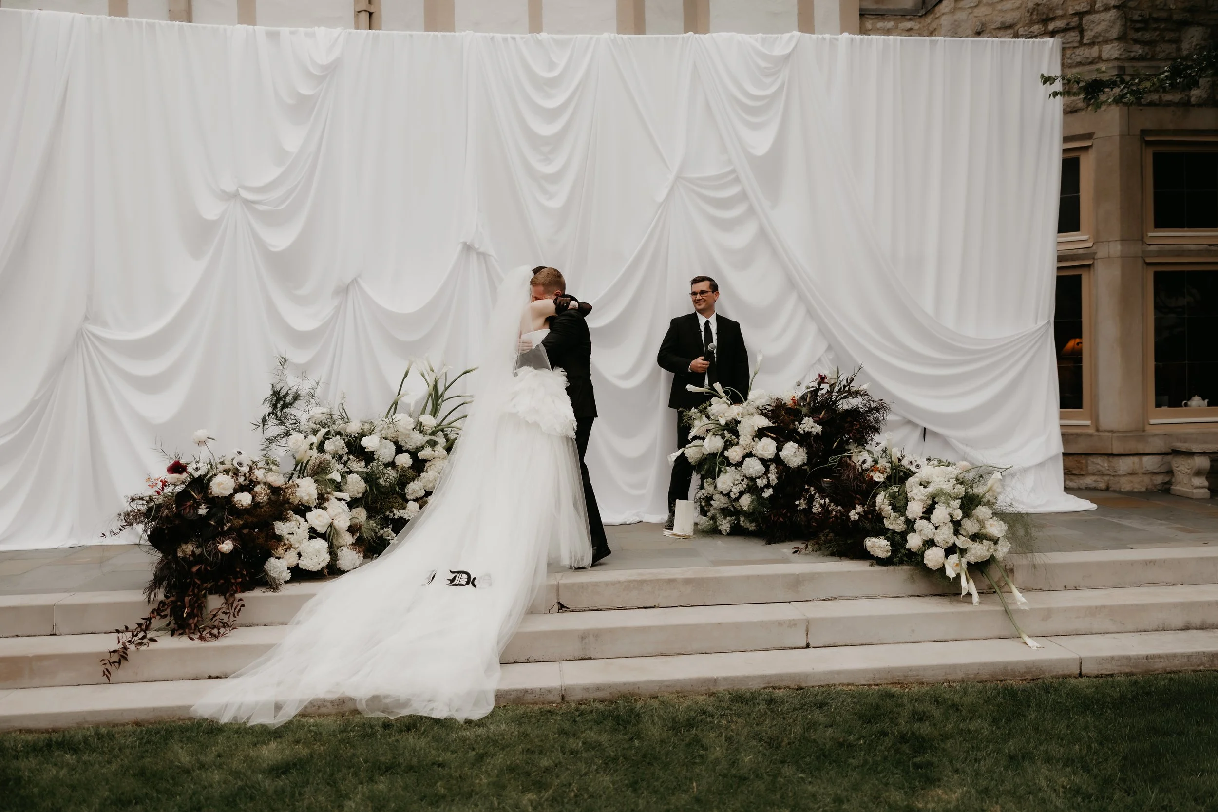 Bride embracing groom during her outdoor wedding ceremony beside white draping and organic floral arrangements, photographed by Julia Brown Photo.