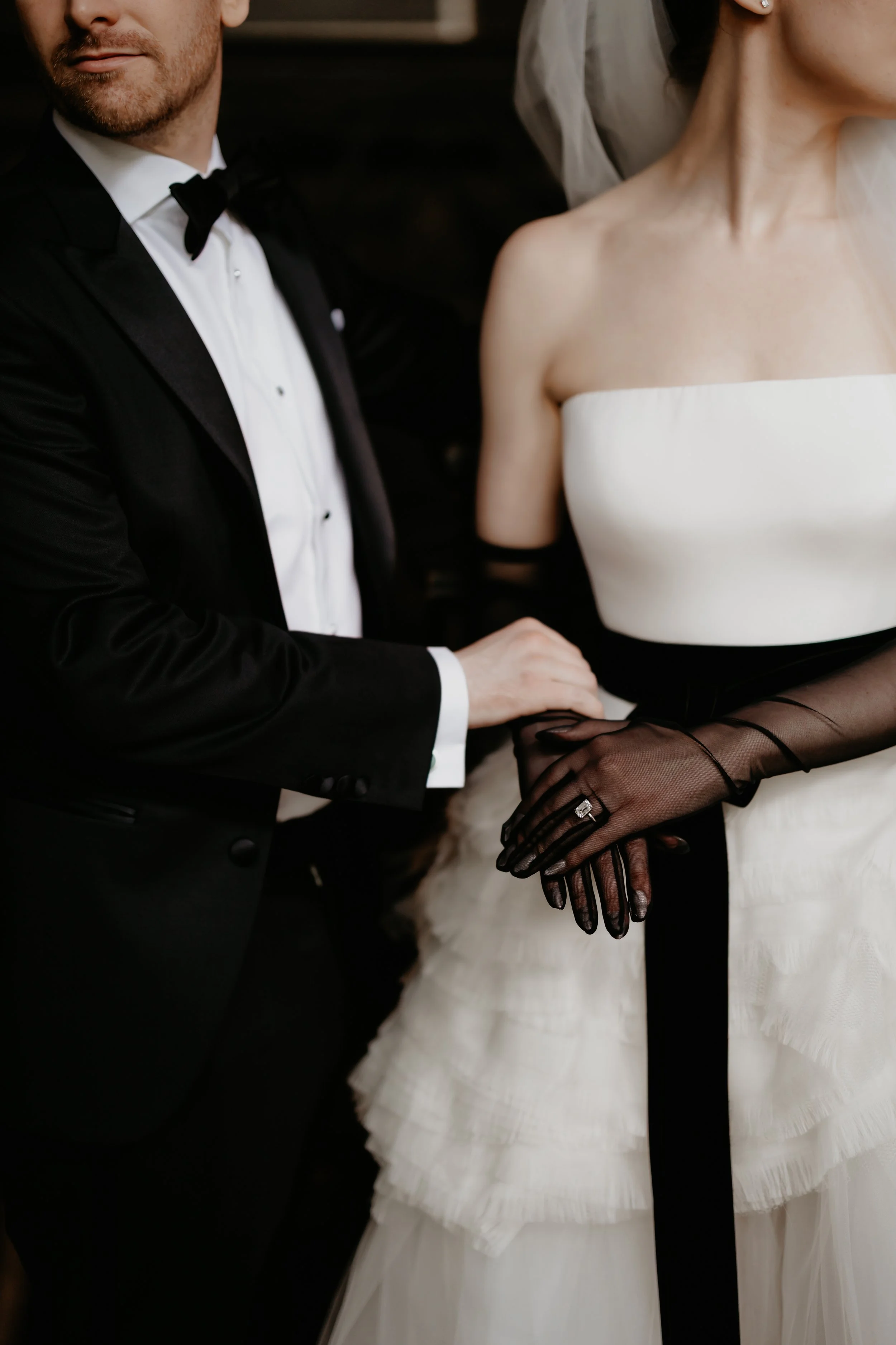 Close-up of the bride’s black opera gloves and black velvet sash beside the groom in a classic black tuxedo, photographed by Julia Brown Photo.